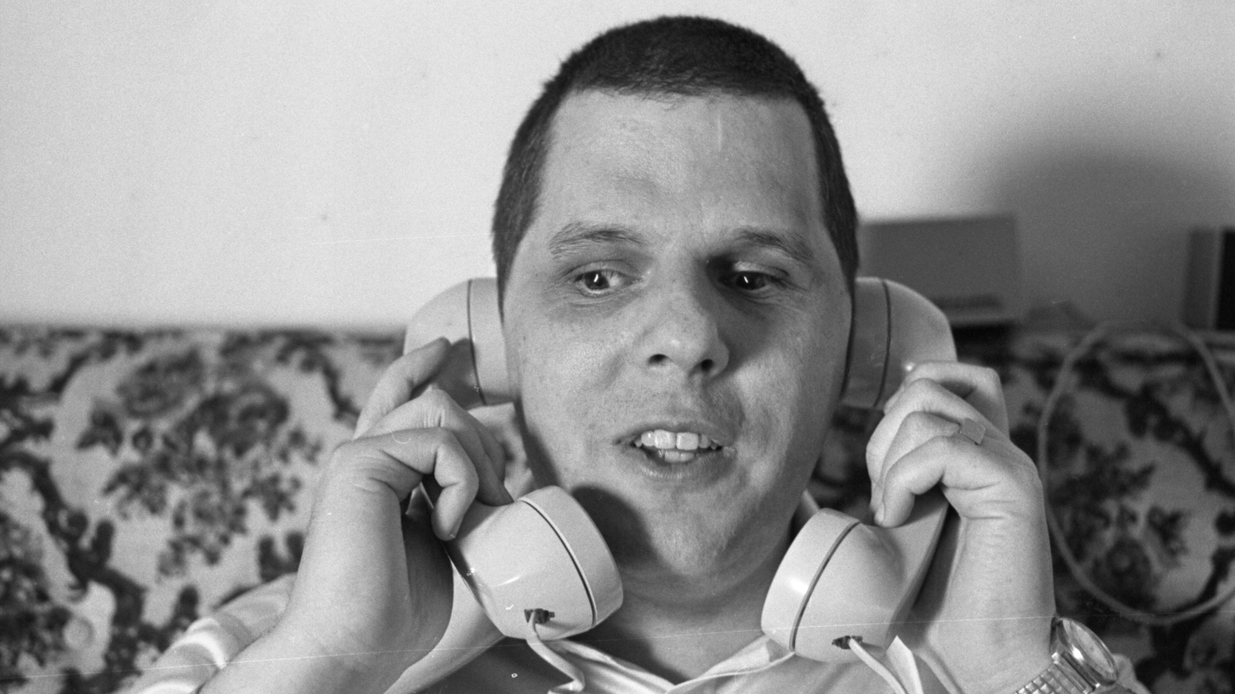 A close-up of a man holding a wired landline telephone to each of his ears. The photo is black and white.