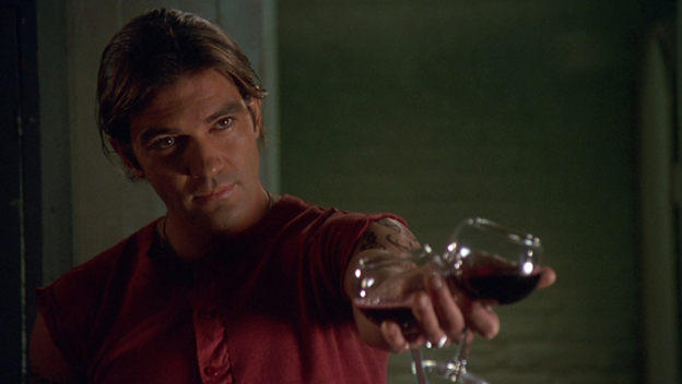 Antonio Banderas faces the camera with his arm extended, holding two glasses of red wine in his hand.