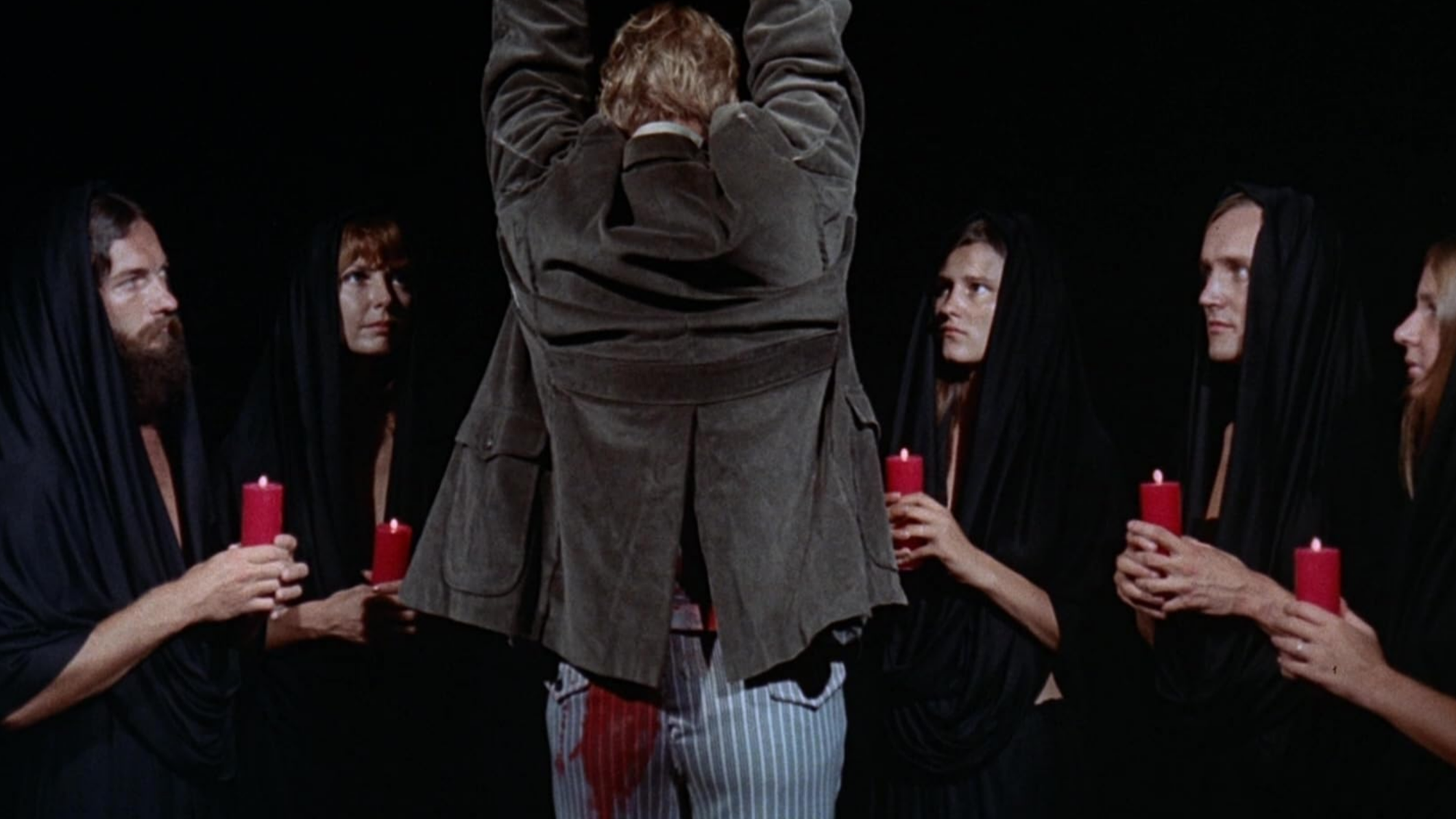 A man held up by his wrists, back to the camera, blood on his pants, surrounded by figures in black hoods holding candles.