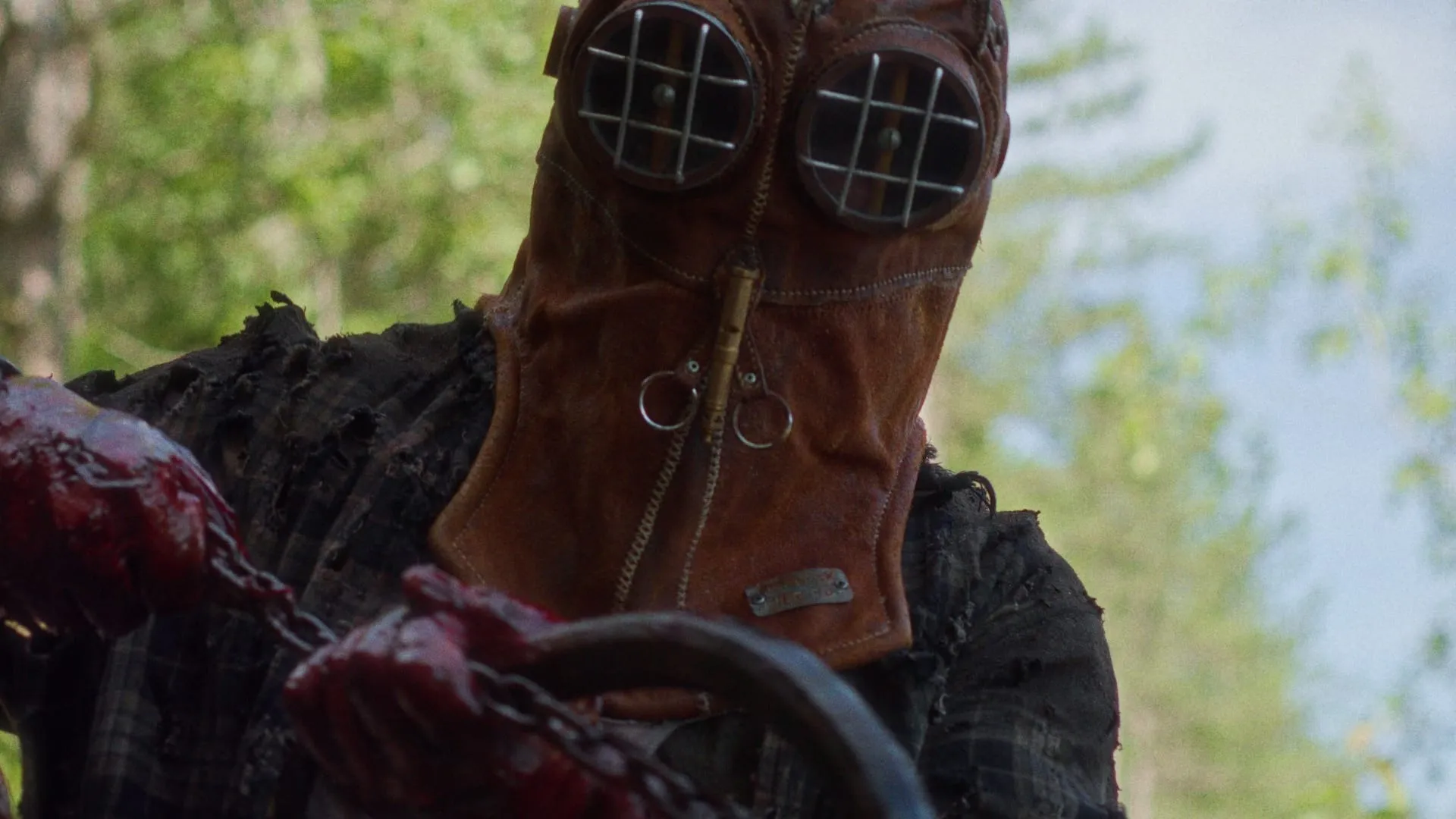 A figure wearing a patchwork mask and a ragged flannel shirt stares into the camera, holding a rusty chain in its bloody hands.