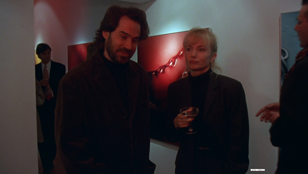 A man with dark hair and a light beard stands next to a blonde white woman holding a coup glass in an art gallery.