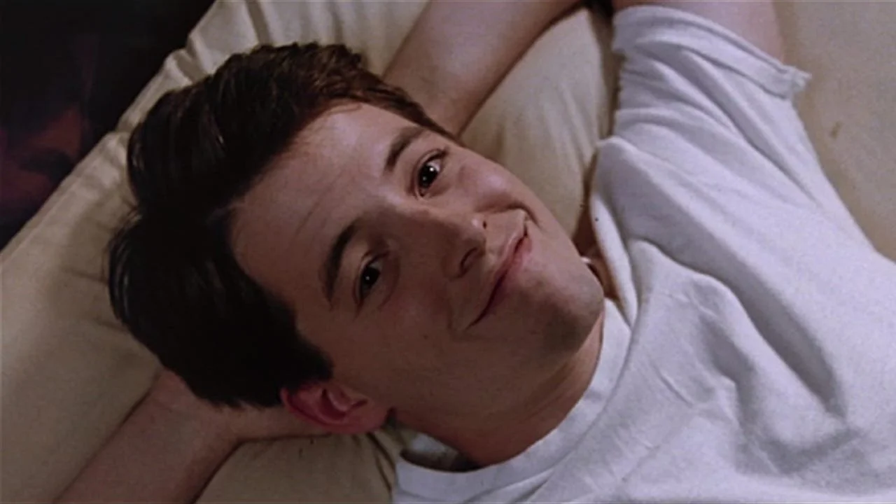 Matthew Broderick relaxes in bed with his hands behind his head and smirks directly at the camera.