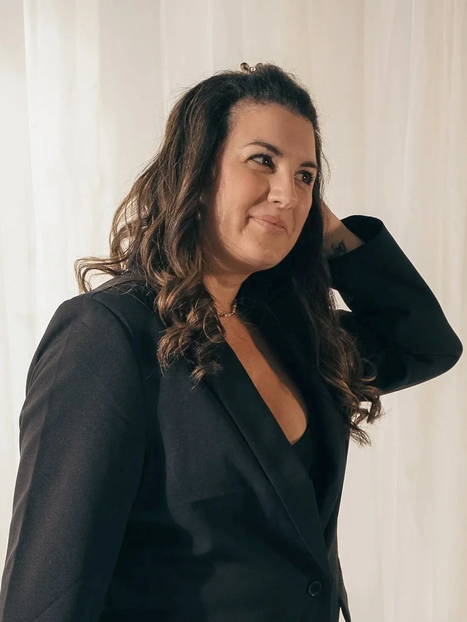 A woman with long, wavy brown hair, wearing a black blazer, standing indoors with a neutral expression and one hand behind her head.