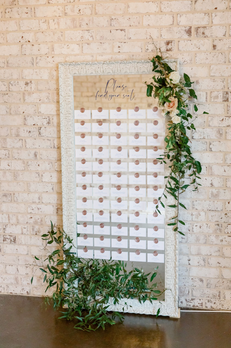 A decorative seating chart board with white cards and copper seals on a full-length mirror, surrounded by green foliage and flowers, against an exposed brick wall.