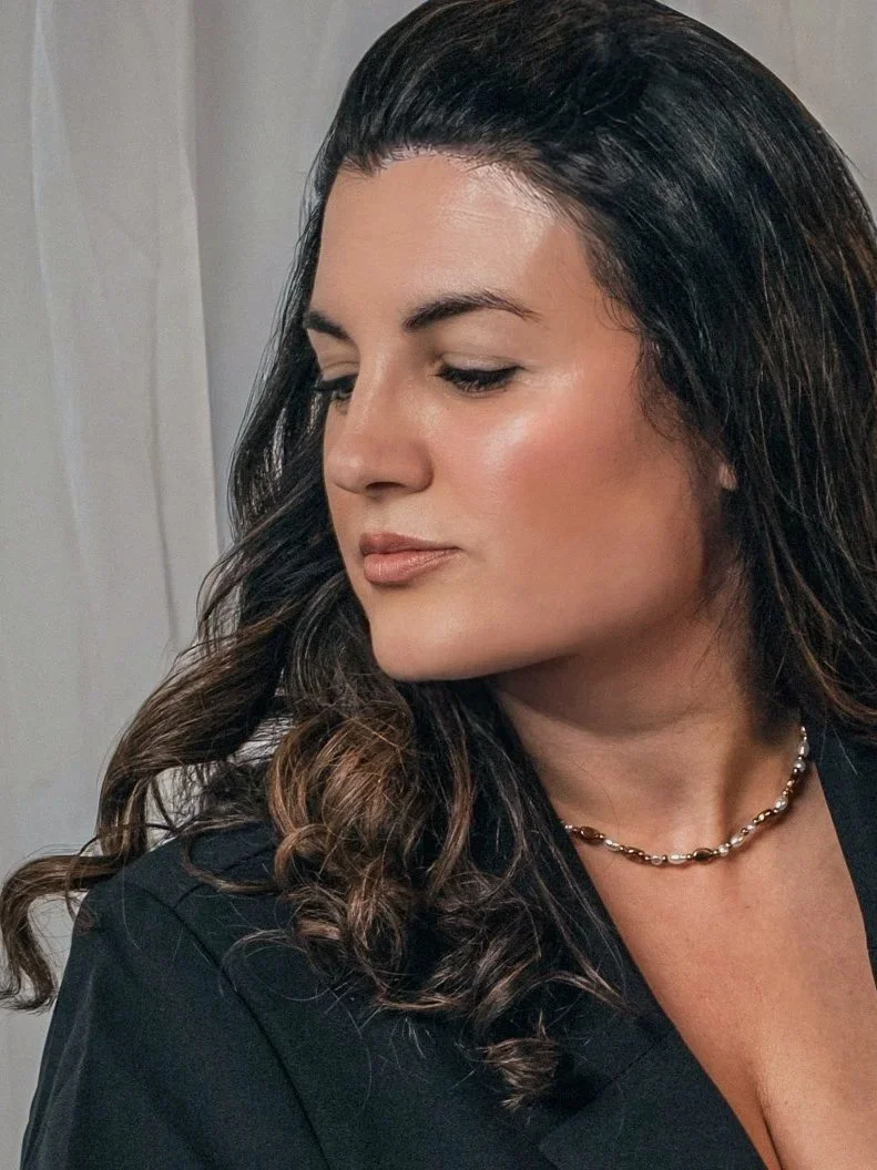 A woman with dark brown, curled hair and light makeup, wearing a black top and a gold chain necklace, looking down and to the side.