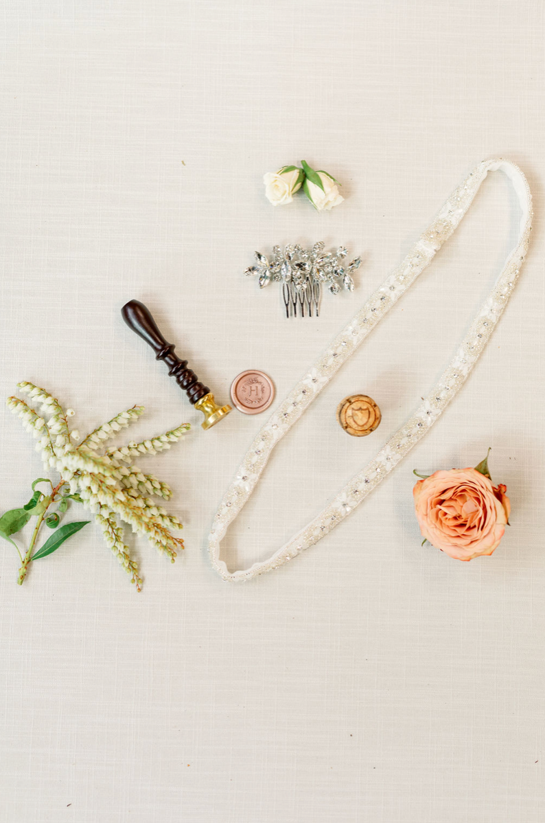 Flat lay of wedding accessories including a flower, hair comb, pearl headband, wax seal, seal stamp, and flower arrangement on light-colored fabric background.