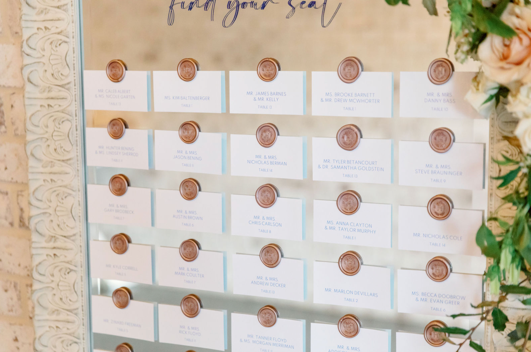 Wedding seating chart with names of guests on white cards attached to a glass board with copper wax seals, bordered by decorative cream-colored molding and floral arrangements on the side.