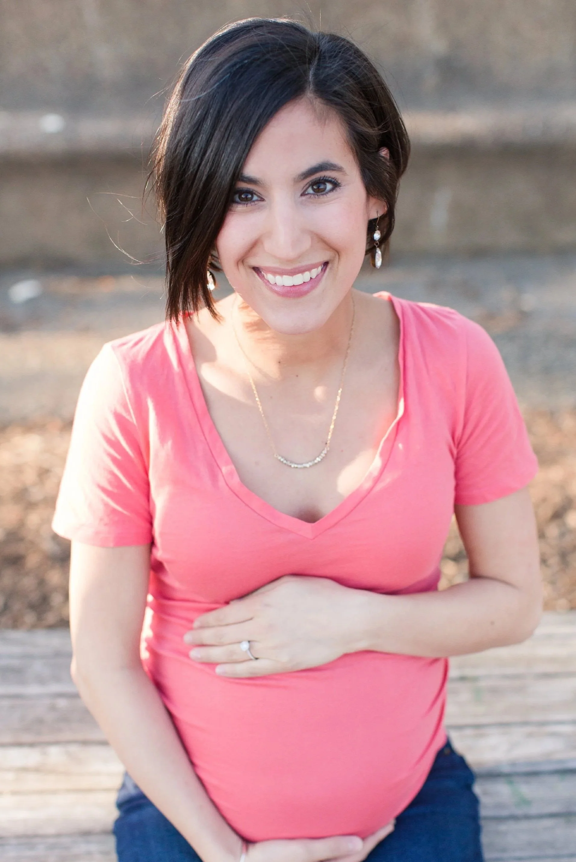 A woman with short dark hair and earrings, smiling while sitting on a wooden surface outdoors, wearing a pink top and dark pants.