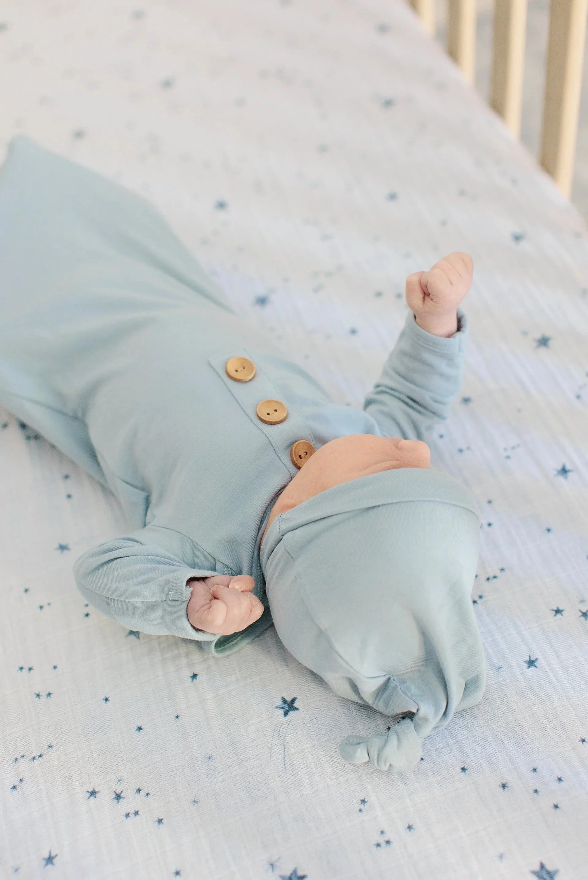 A baby lying on a white blanket with blue stars, wearing a light blue outfit with buttons on the sleeve, and showing a clenched fist.