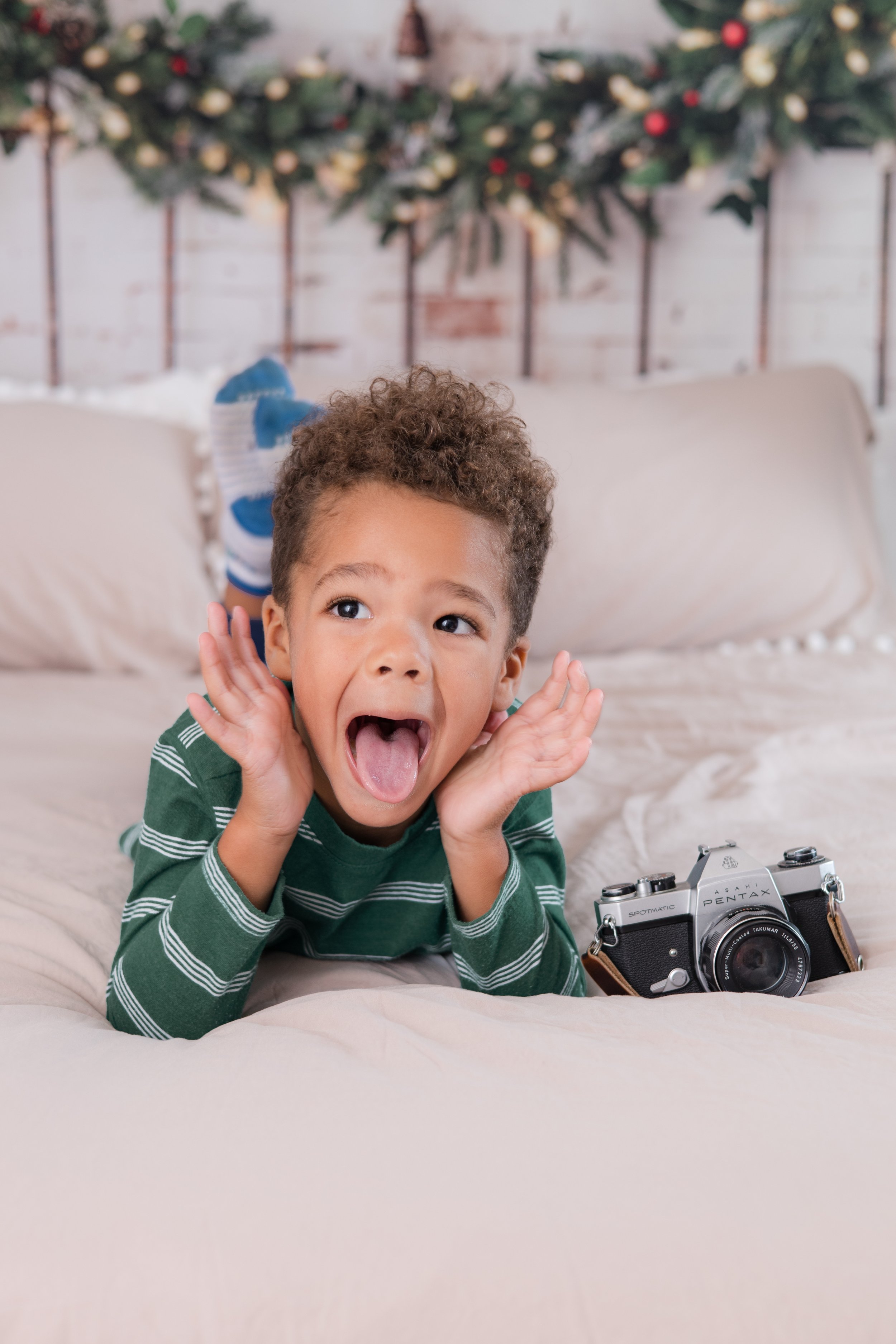 A young boy with curly hair and a striped green shirt is lying on a bed with a vintage camera next to him. He is making a playful face with his mouth open and hands near his ears. In the background, there is a decorated Christmas tree with ornaments and a white brick wall.