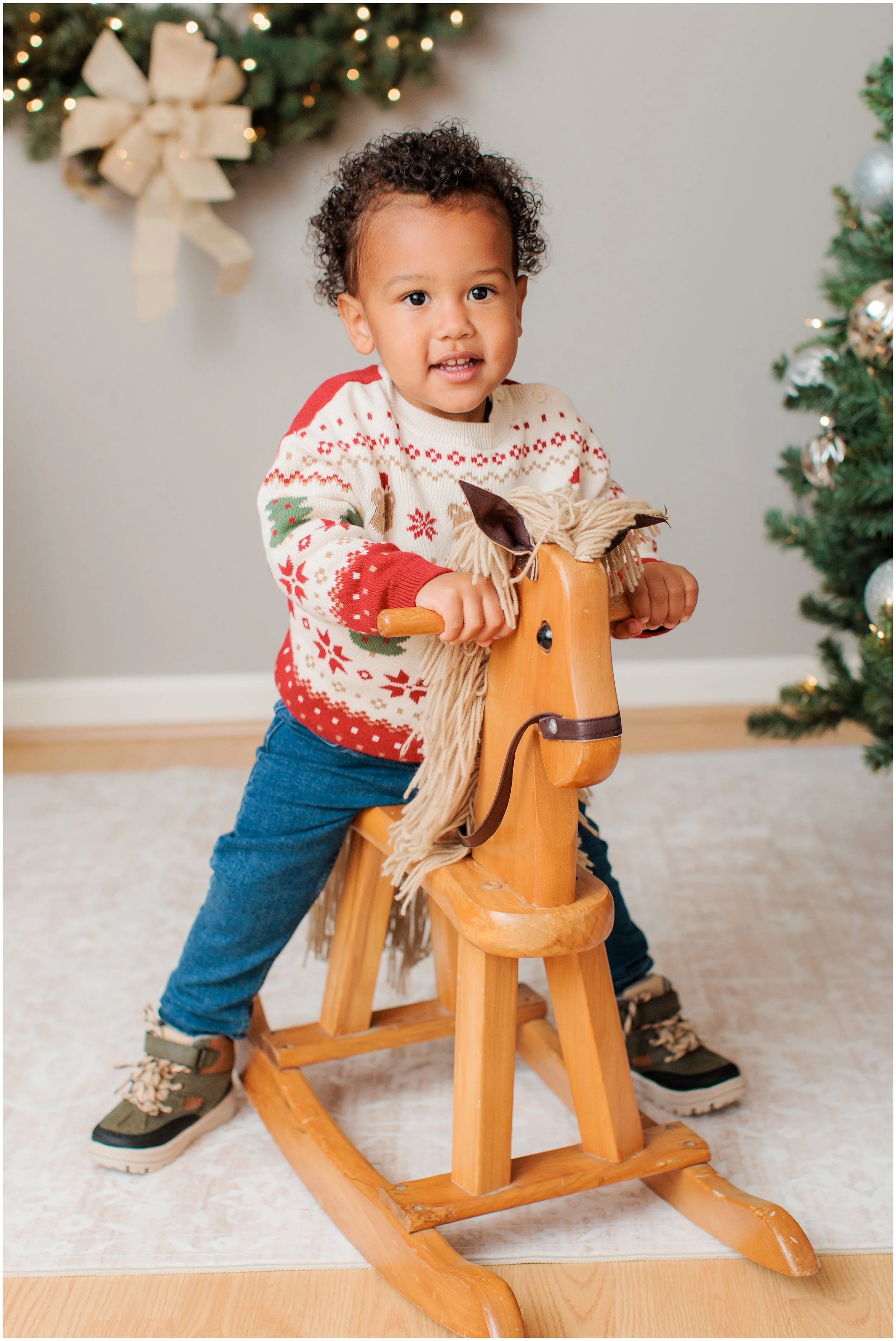 A young child with curly hair riding a wooden rocking horse in a room decorated for Christmas with a Christmas tree and a wreath with a large bow in the background.