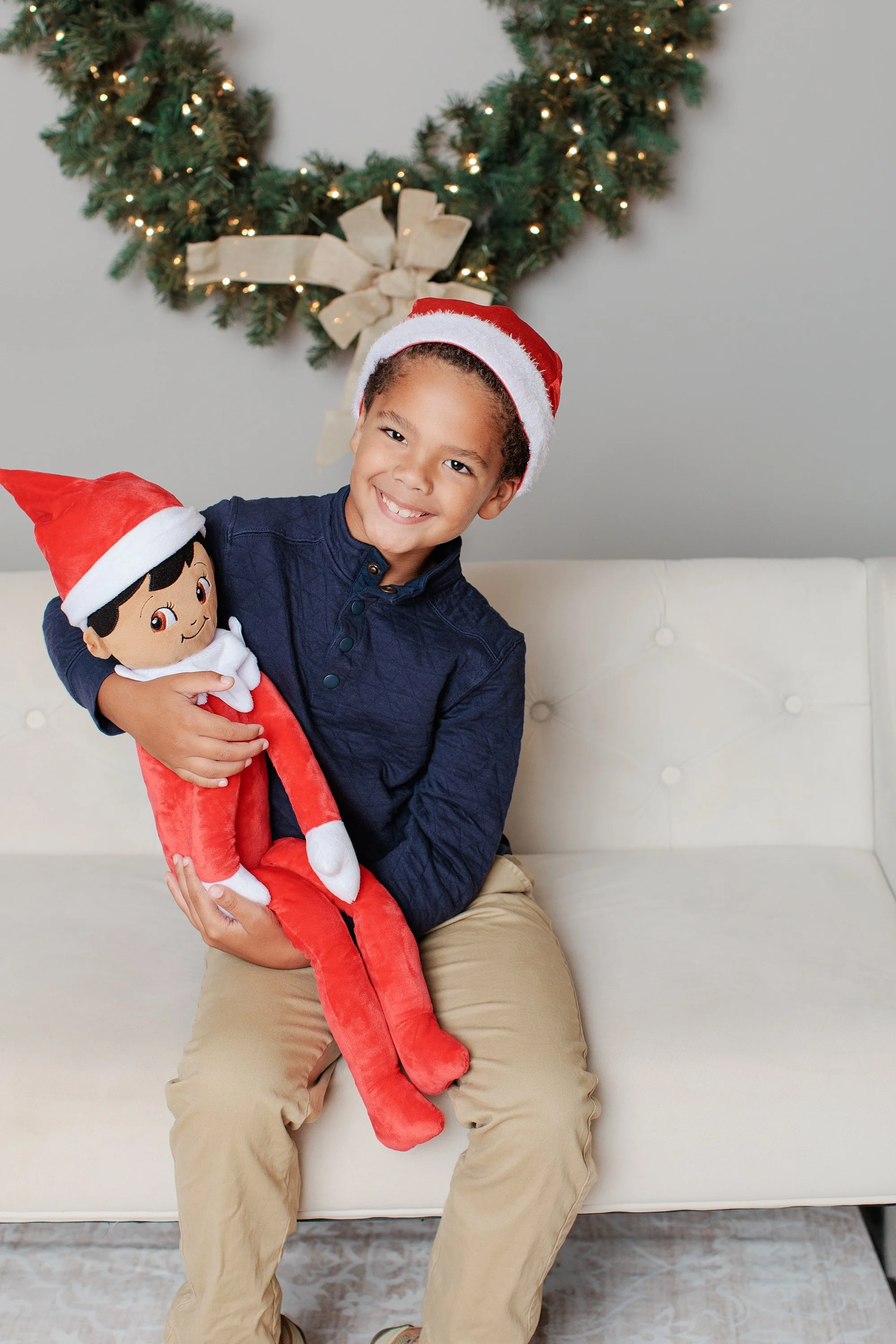 Boy wearing a Santa hat and holding a plush Christmas elf on a cream sofa.