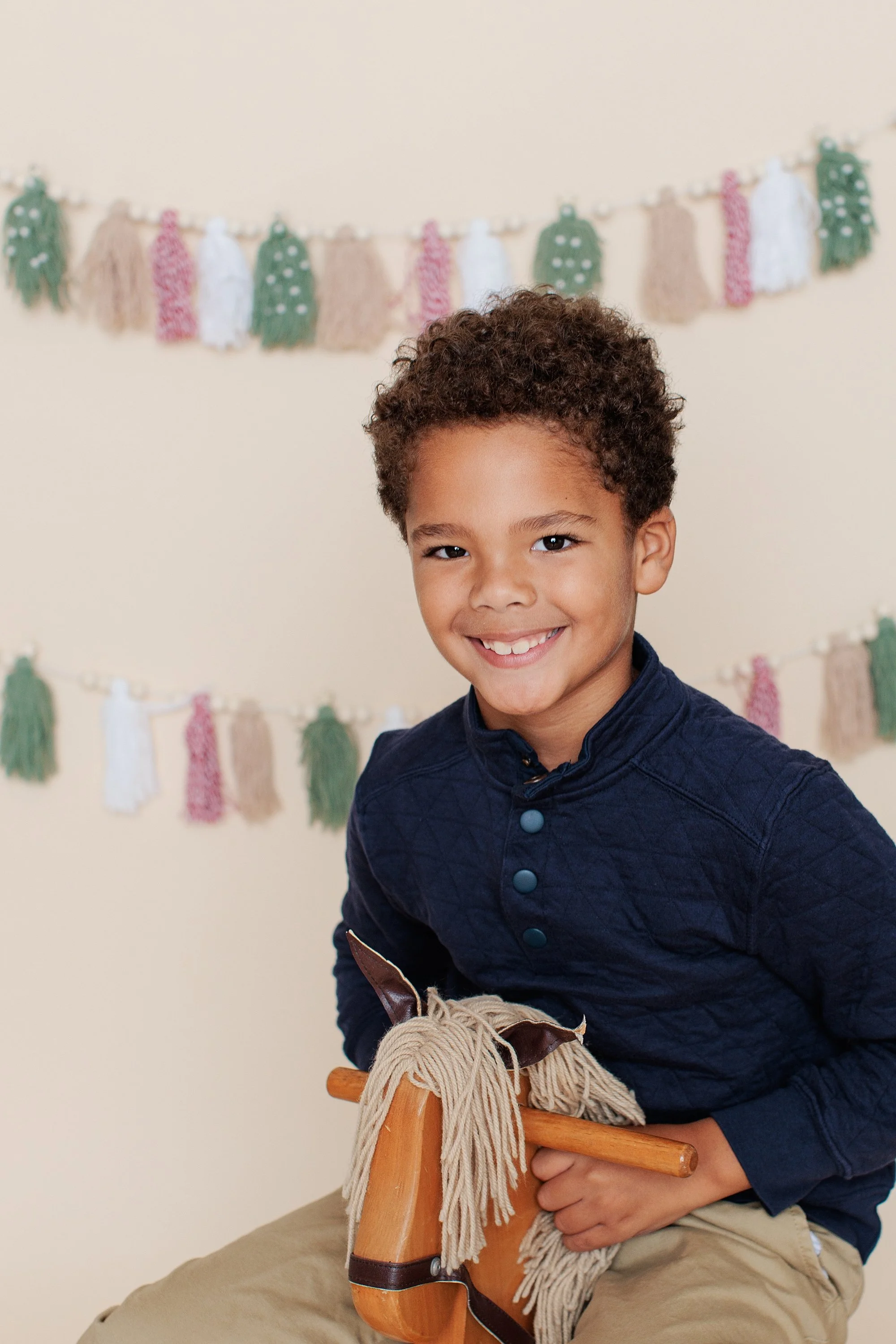 Boy turned slightly to the side on a wooden rocking horse against a cream tassel backdrop.