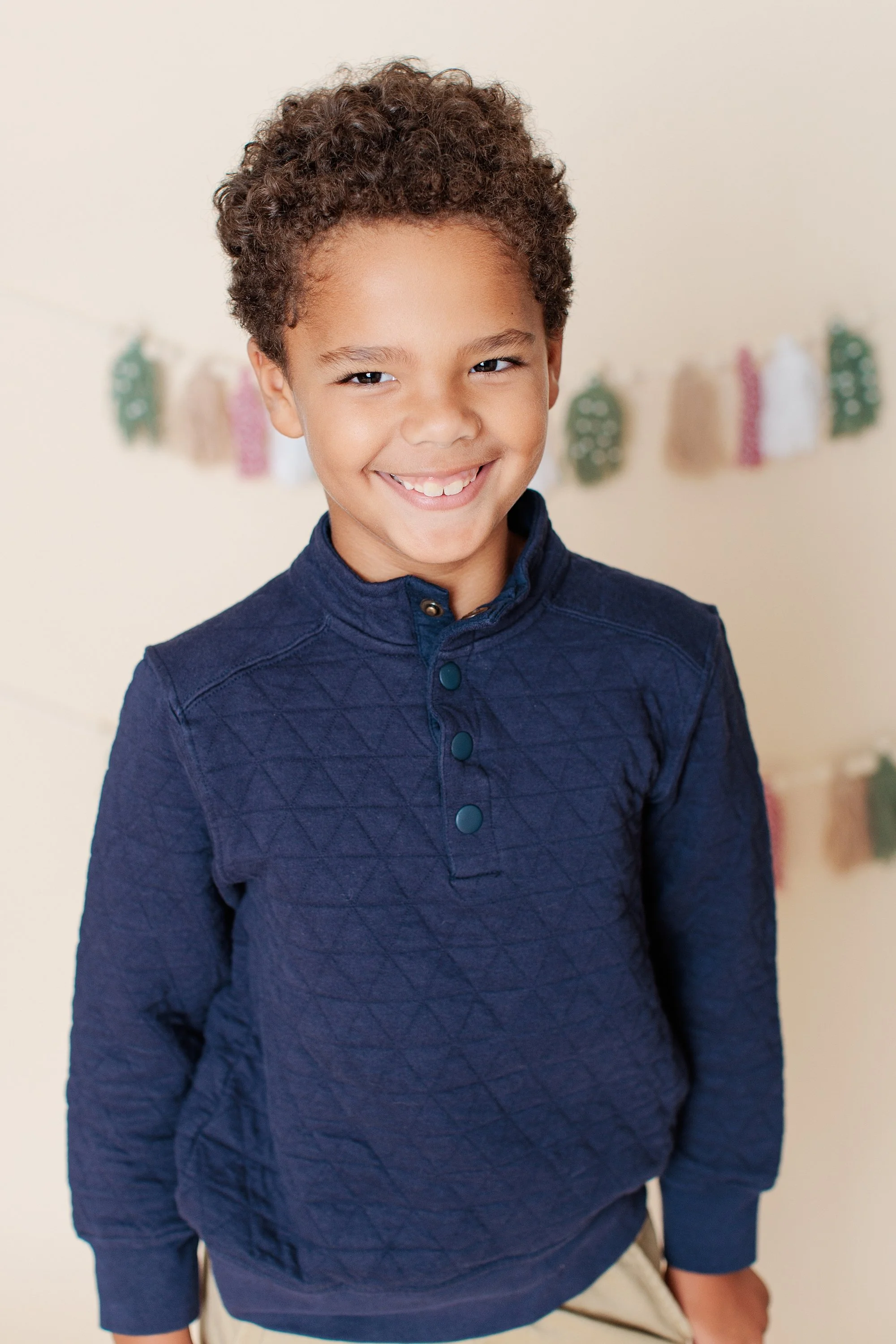 Smiling portrait of a boy in a textured navy top with muted holiday tassels behind him.