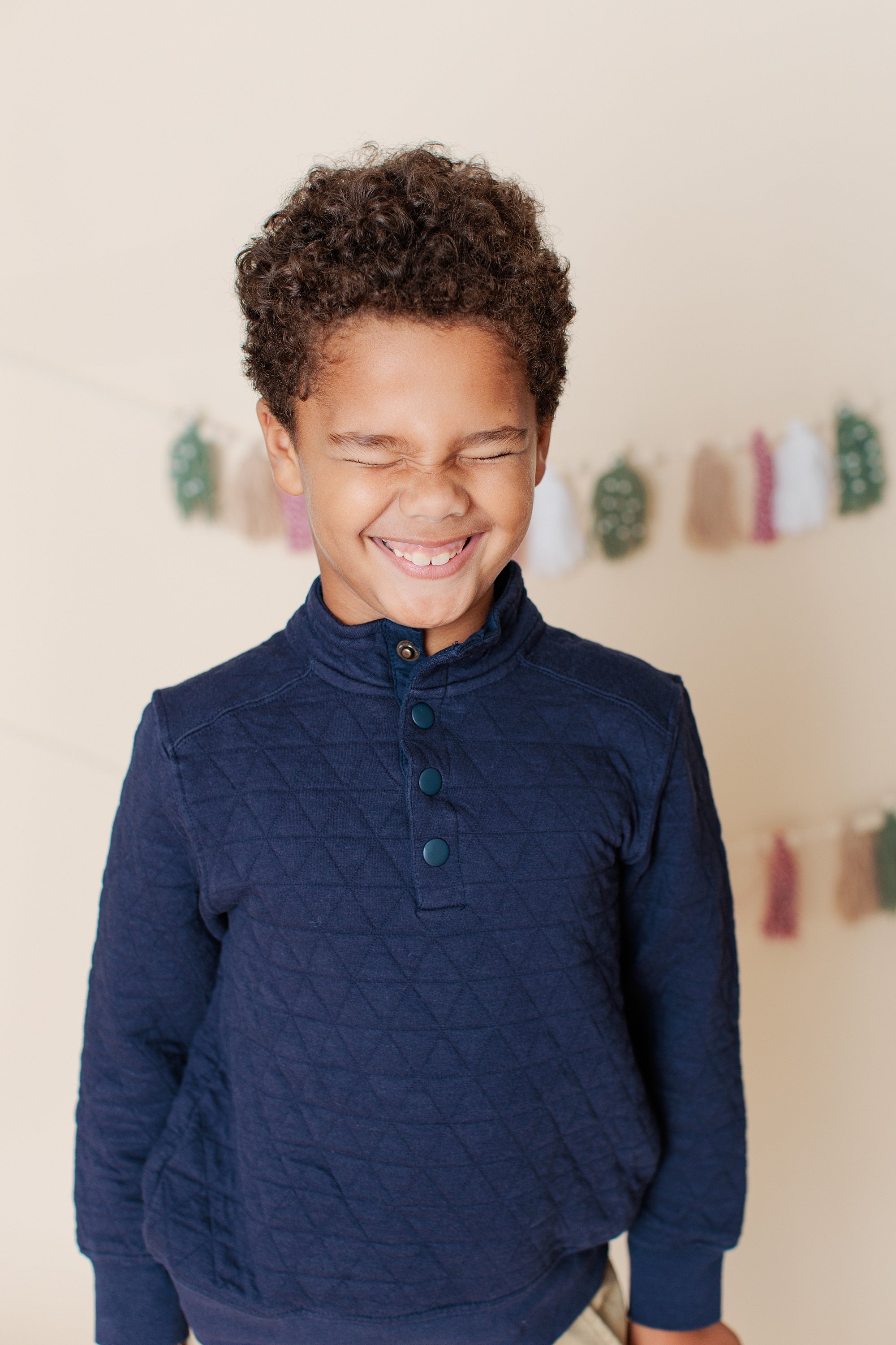 Boy grinning with his eyes squeezed shut in front of a holiday tassel backdrop.