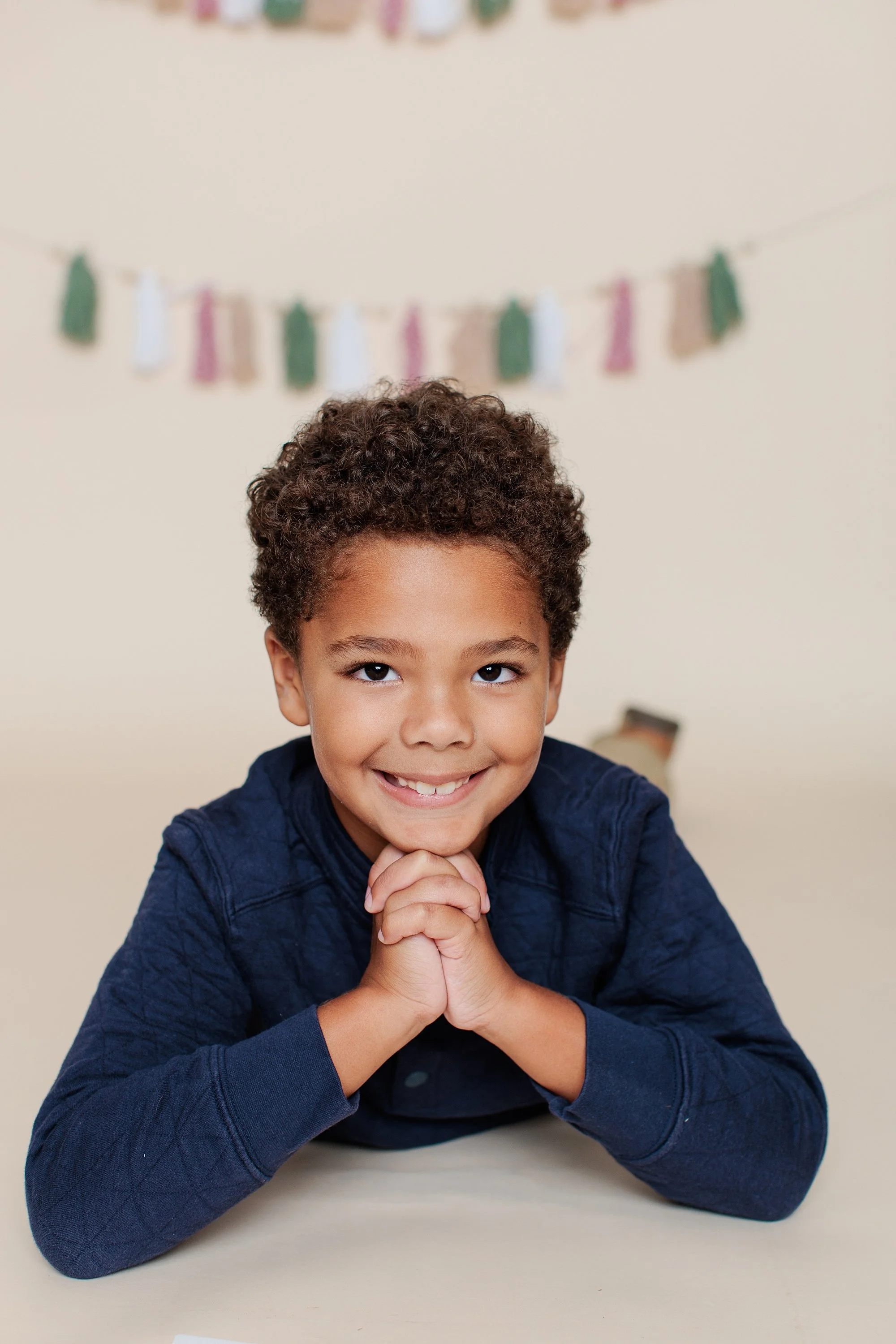 Boy smiling at the camera with his chin resting on clasped hands against a cream holiday backdrop.