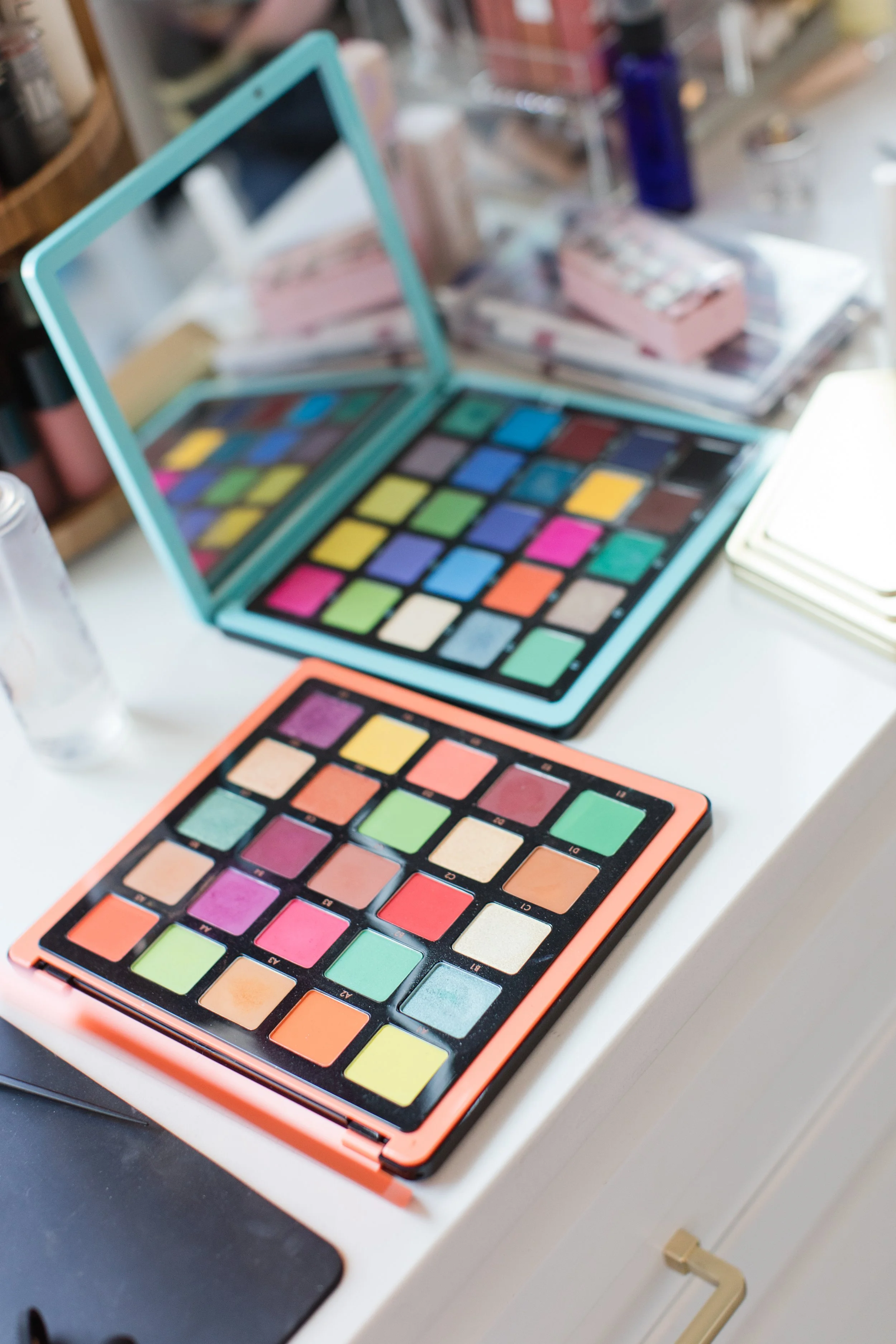 Two colorful eyeshadow palettes with a variety of shades are placed on a white surface, with makeup items and bottles blurred in the background.