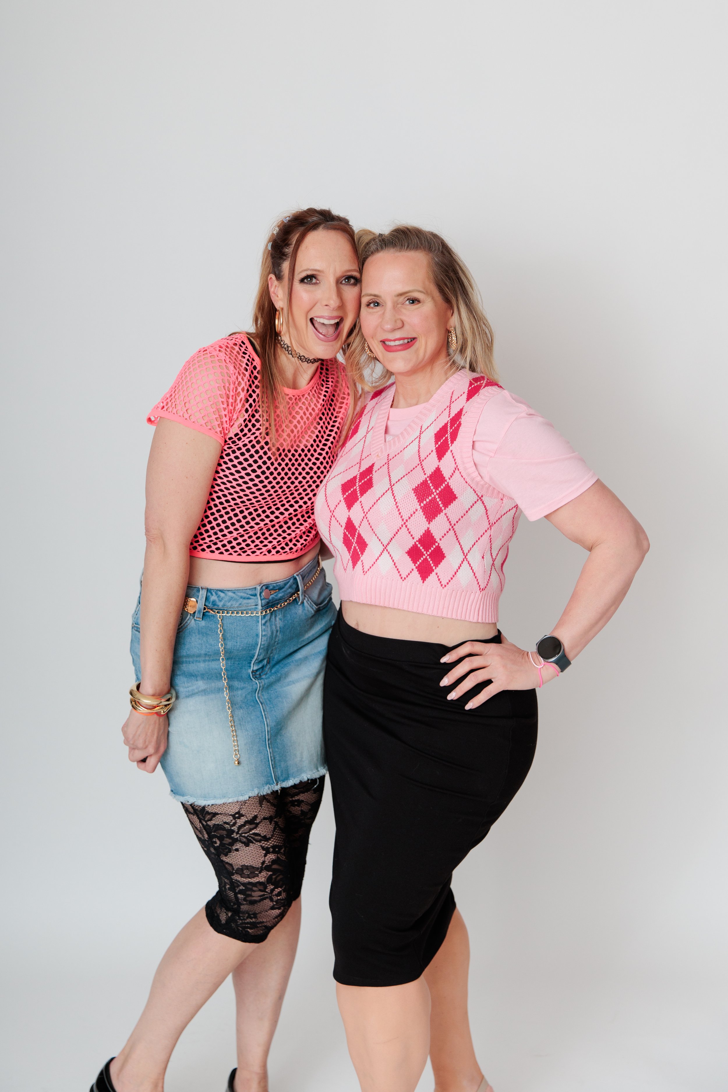 Two women standing close together, smiling, against a plain white background. One wears a pink mesh top and denim skirt, the other a pink argyle sweater vest over a pink T-shirt and black skirt.