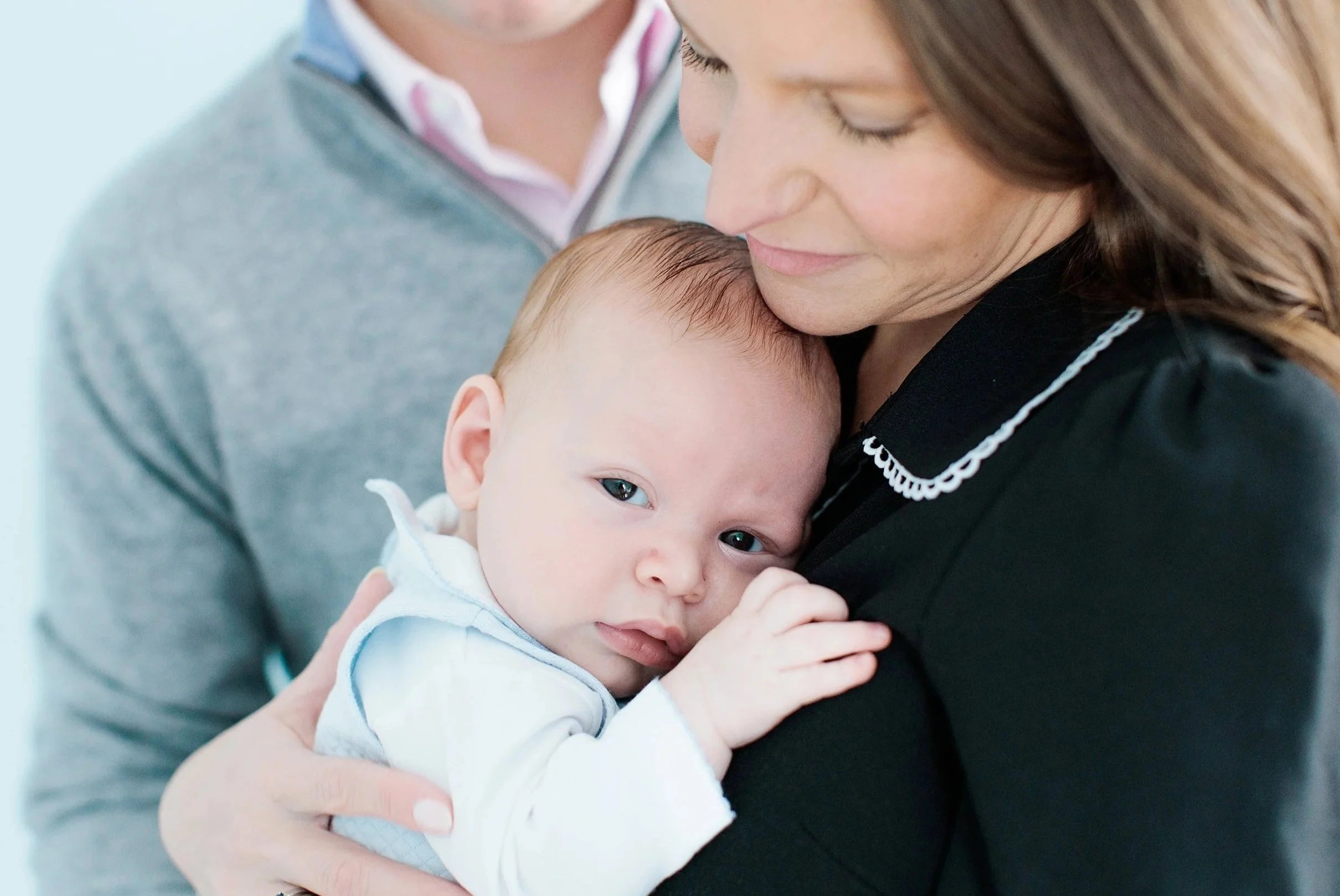 A woman is holding a baby close to her chest, with their faces touching. The woman has brown hair and is smiling gently, while the baby looks content and has light brown hair. The background is plain and softly lit.