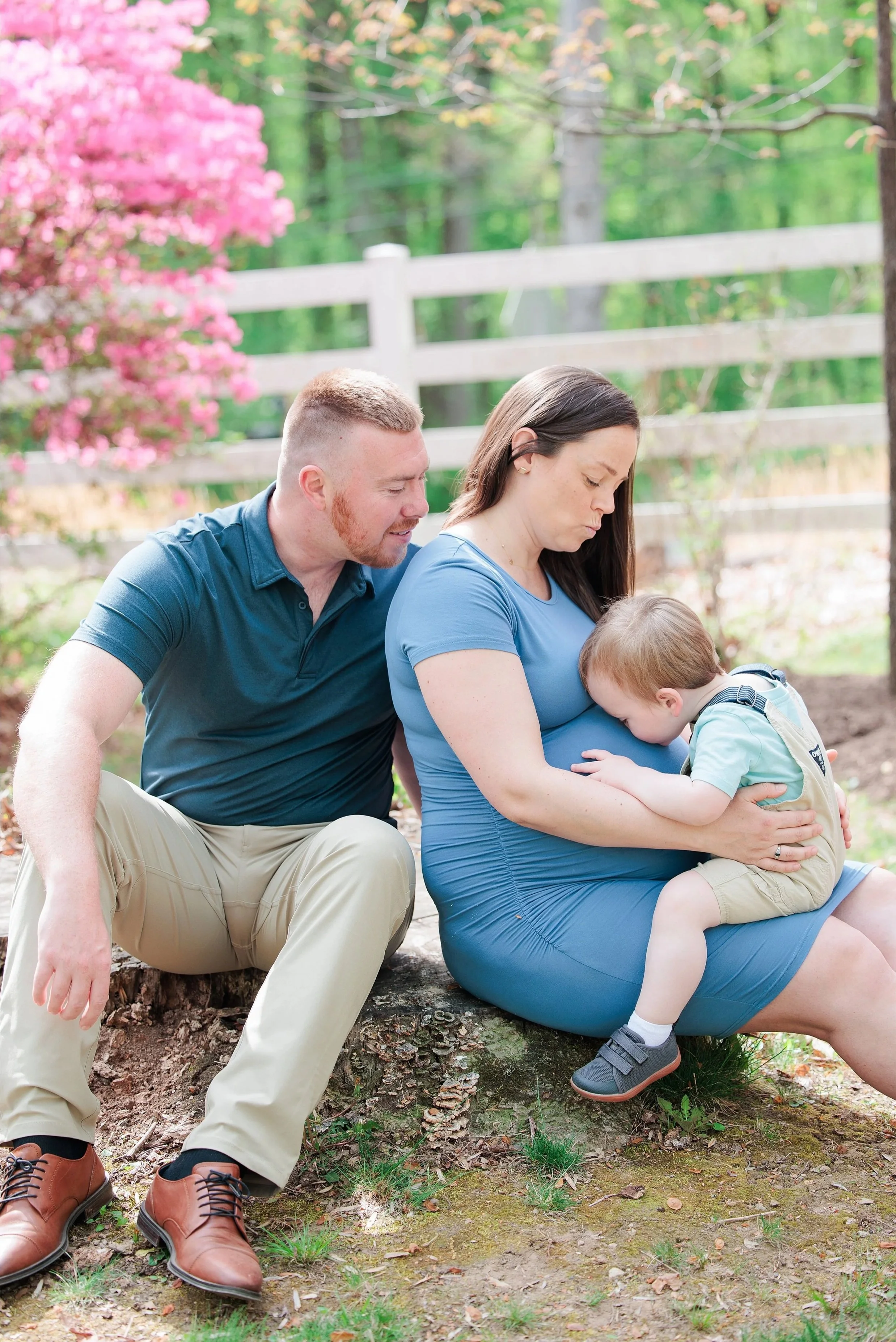 A man and woman with a young child outdoors. The man crouches next to the woman, who is sitting on a log, as the child leans into her. They are in a wooded area with trees, a pink-flowered bush, and a white fence in the background.