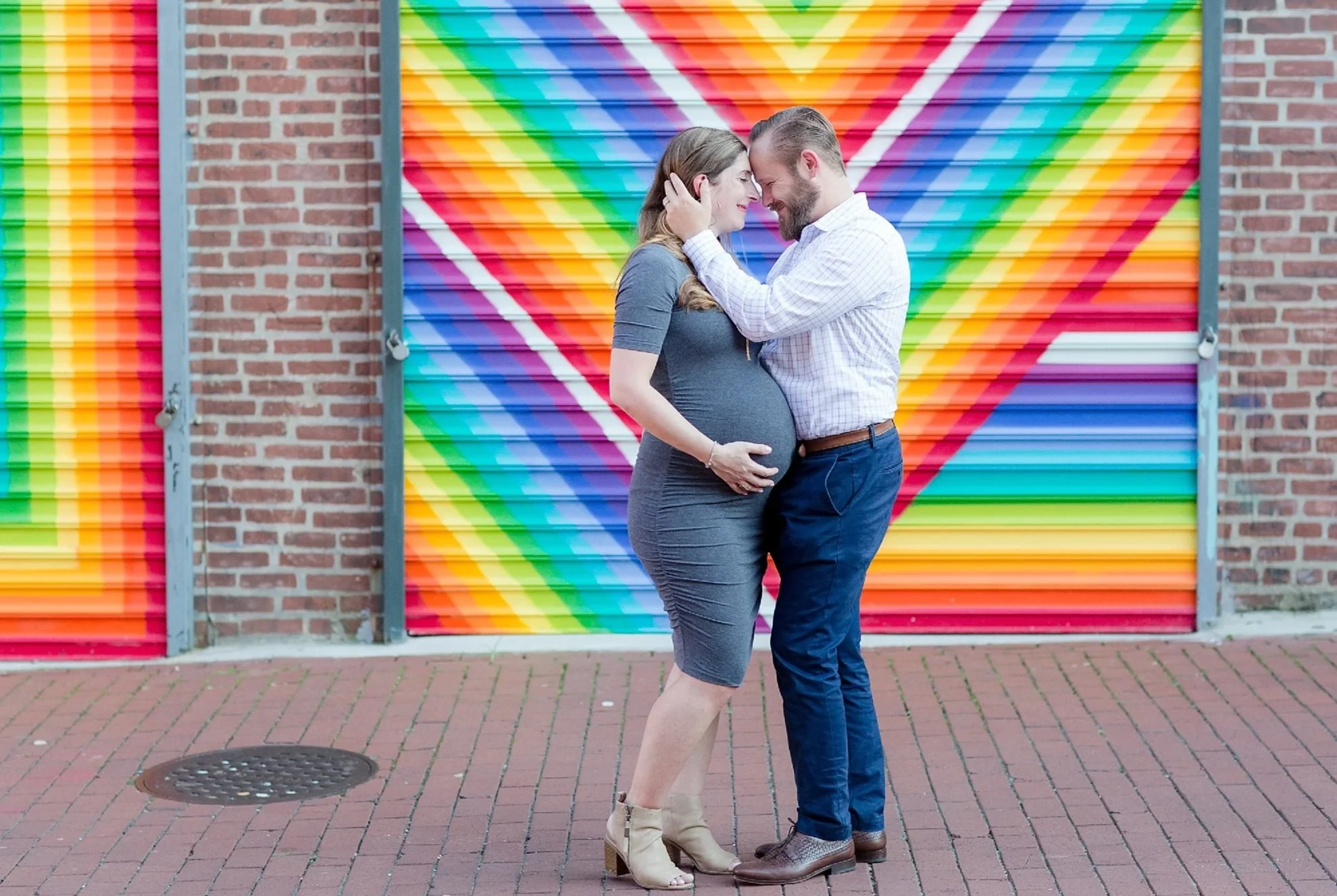 A couple stands close together touching foreheads, with their eyes closed, in front of a colorful rainbow mural painted on a metal garage door and brick wall. The woman is pregnant and wearing a gray dress and beige high heels, while the man is wearing a white striped shirt and dark pants.