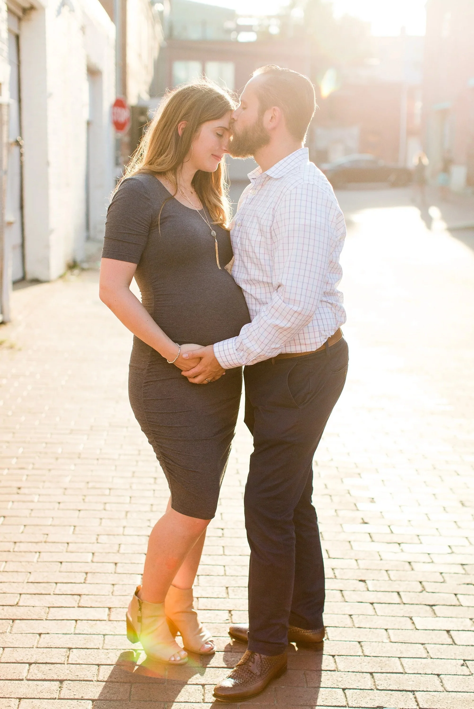 A man and woman, whose bodies are touching, stand on a sunlit street holding hands, with the man kissing the woman's forehead. The woman is pregnant, wearing a dark dress and beige high heels. The man is dressed in a checkered shirt, dark pants, and dress shoes.