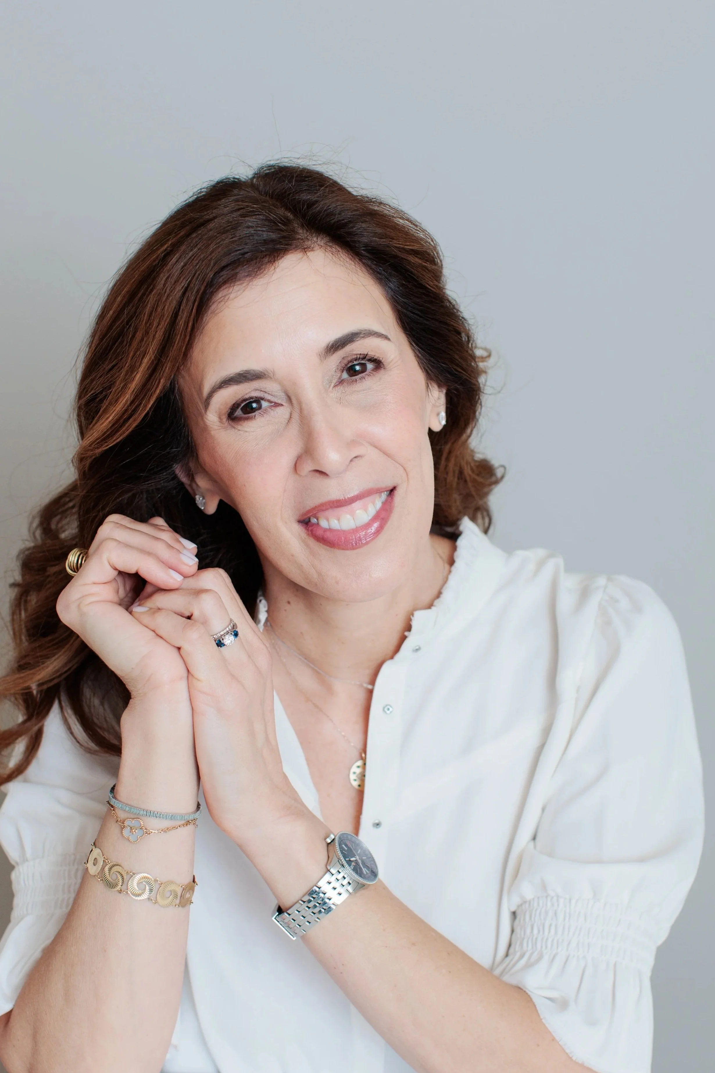 Close-up of a smiling middle-aged woman with long, wavy brown hair, wearing a white blouse, multiple bracelets, a watch, and rings, posing with her hands under her chin against a neutral background.