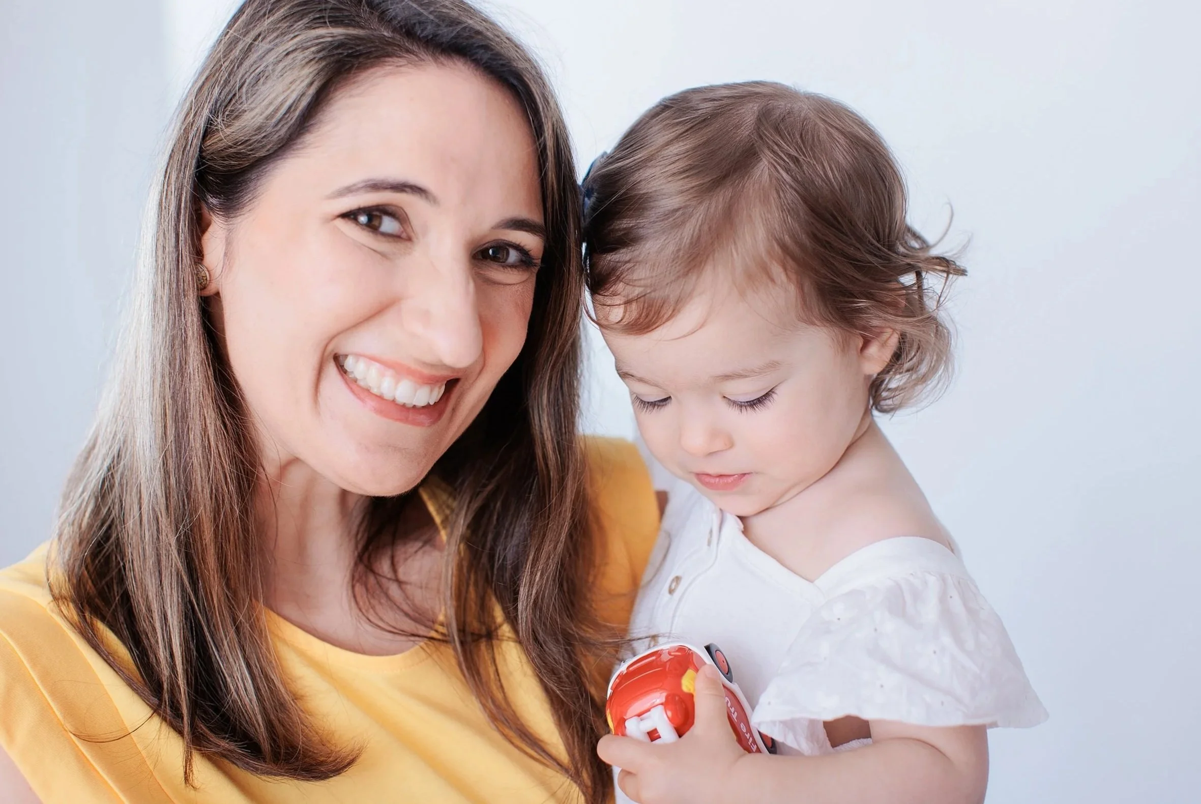 A woman with long brown hair and a young girl with curly hair looking at a toy phone together, both smiling and appearing happy.