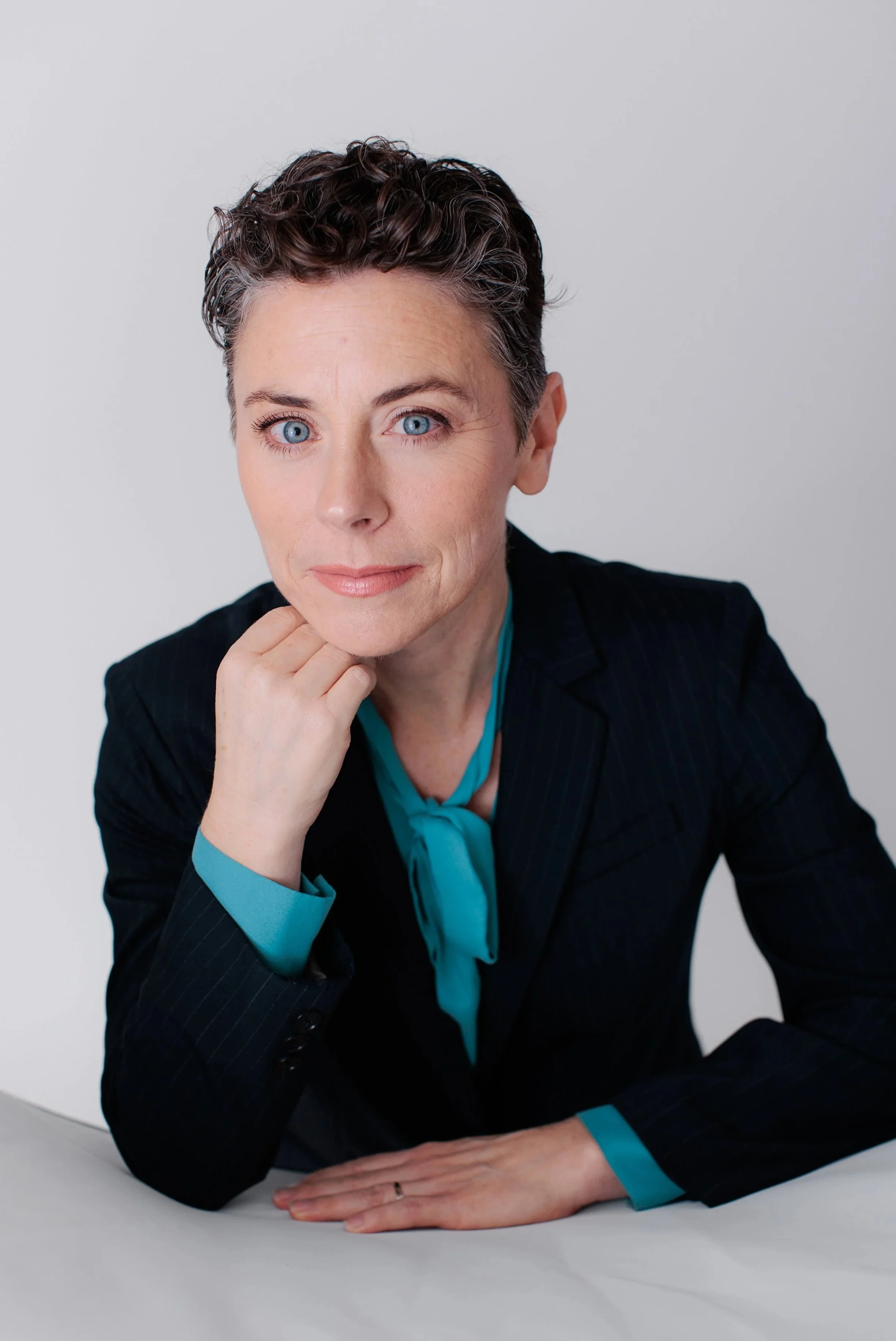 Woman with short curly dark hair, blue eyes, wearing a black pinstripe blazer and a teal shirt, resting her chin on her hand and smiling slightly against a plain white background.
