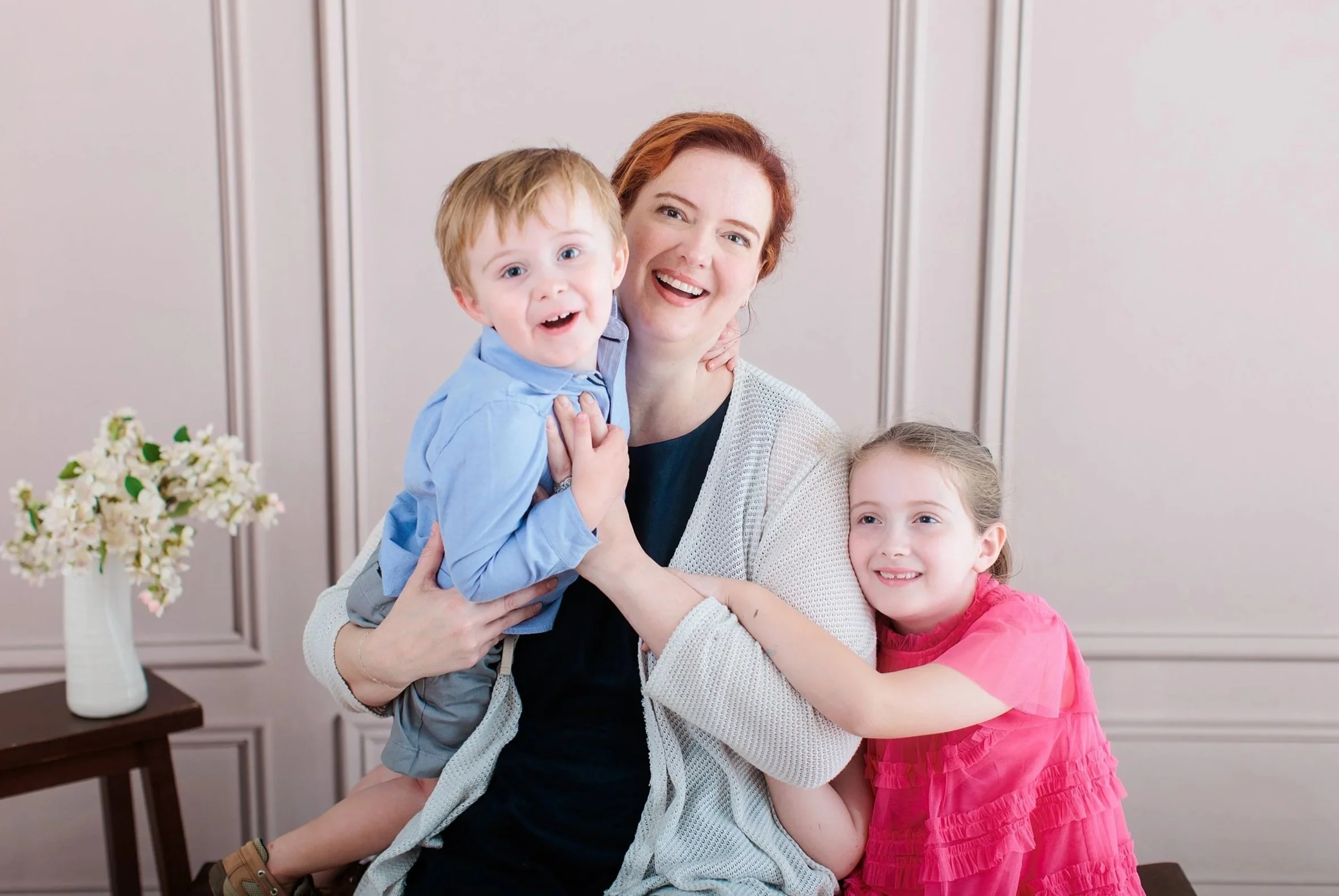 A happy woman with two children hugging her indoors, with a bouquet of flowers on a table to the side.