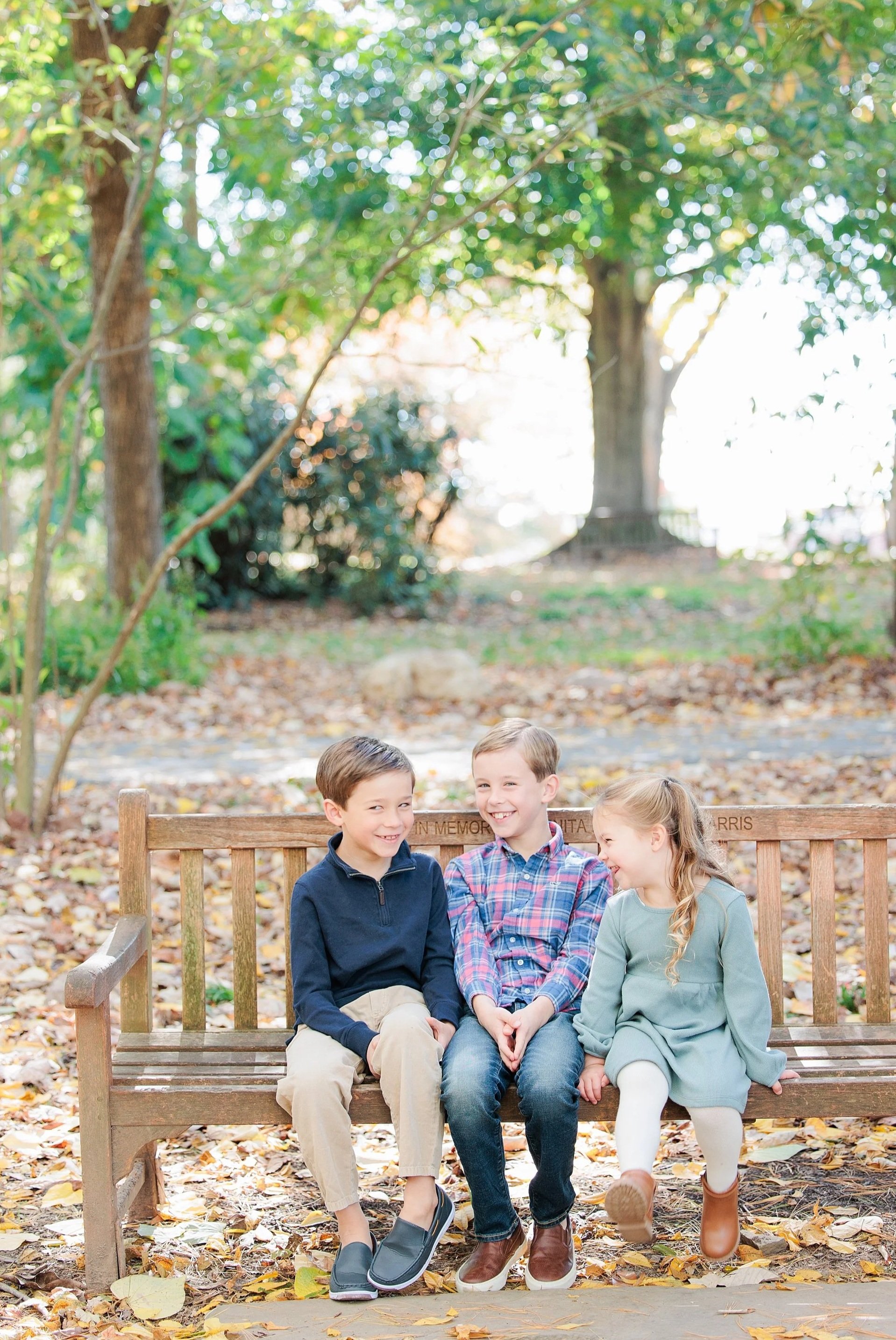 Four children sitting on a wooden park bench with fall leaves on the ground, smiling and talking, with trees in the background.
