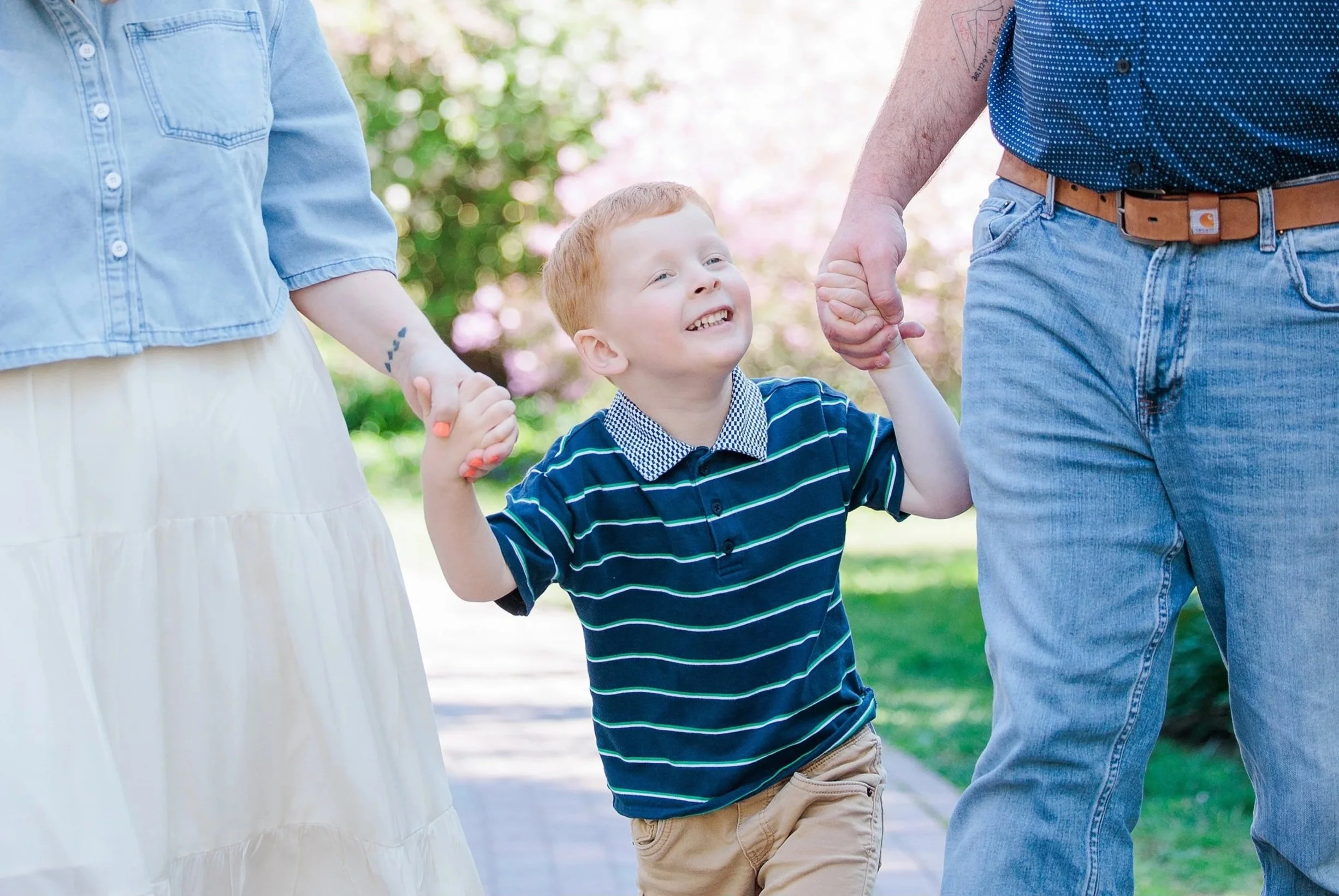 A young boy with red hair smiling and holding hands with two adults, walking outdoors in a park with green trees and sunlight in the background.