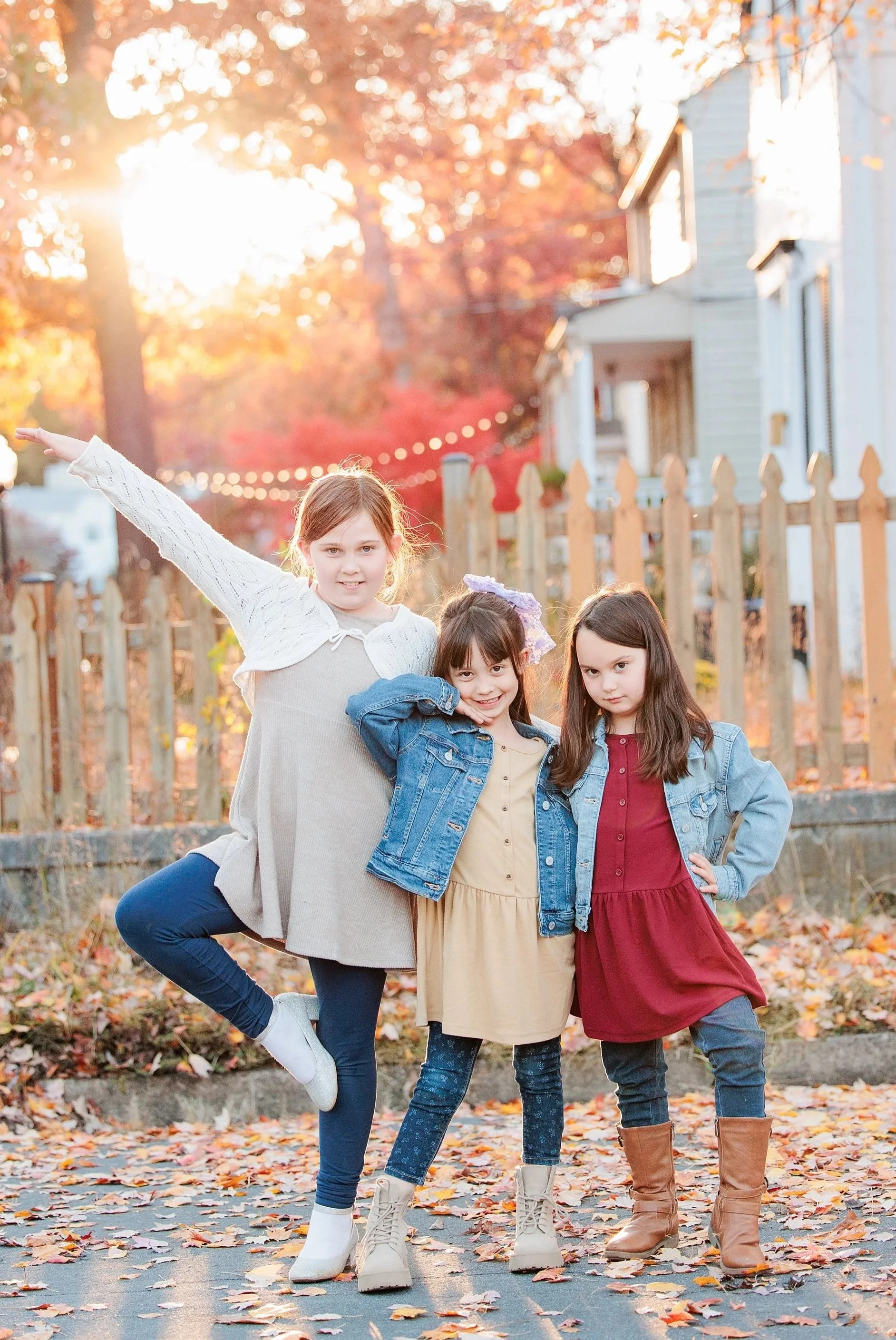 Four young girls standing outdoors on a fall day, surrounded by autumn leaves, with sunlight peeking through trees in the background. They are dressed in casual fall clothing, including boots and denim jackets.