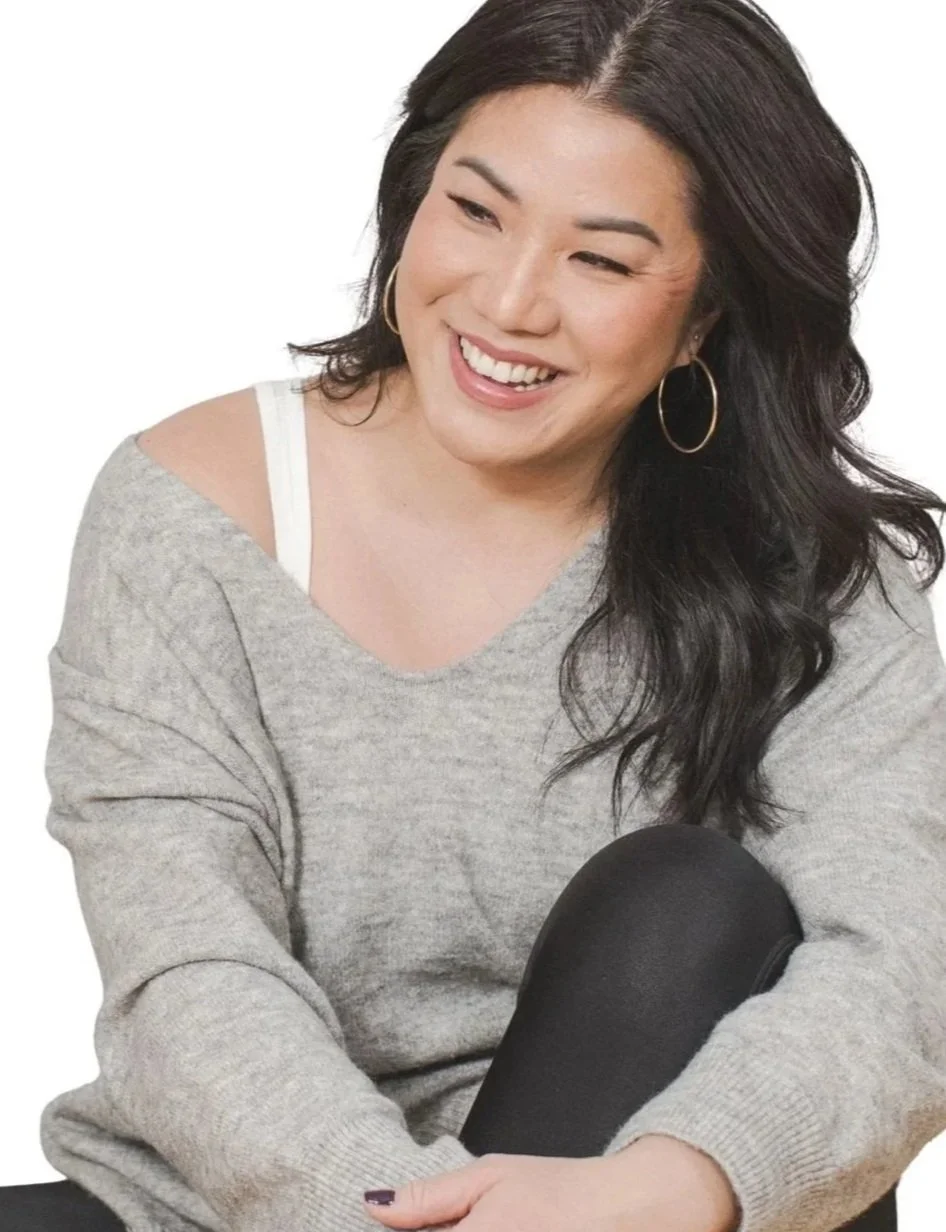 A woman with dark wavy hair, wearing hoop earrings and a gray sweatshirt, smiling and sitting with one knee up.