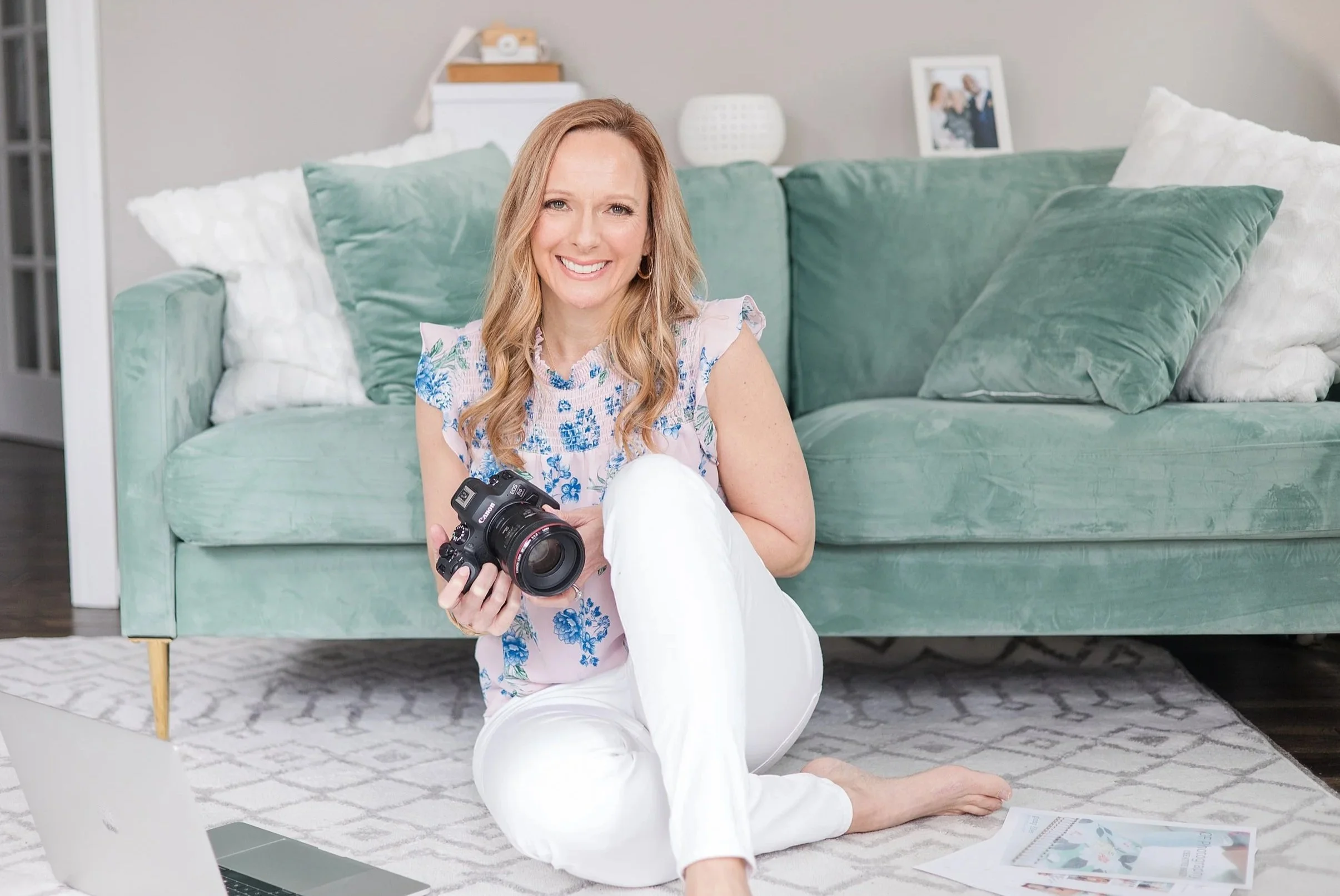 A woman with long, wavy blonde hair smiling and sitting cross-legged on a light-colored rug in front of a mint green sofa, holding a camera in her hands, with photographic prints and a laptop nearby in a cozy living room.