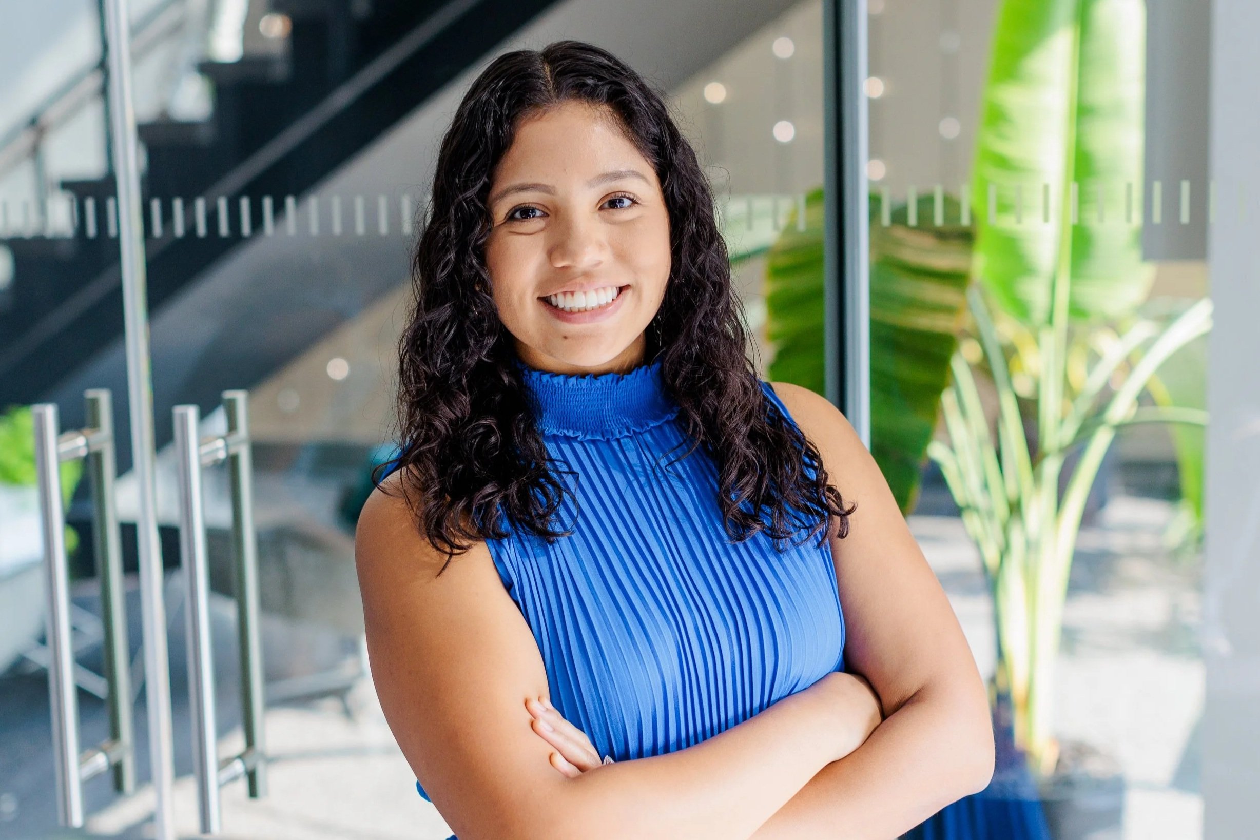 A young woman with curly black hair smiling and standing with arms crossed in front of a glass wall with green plants behind her.