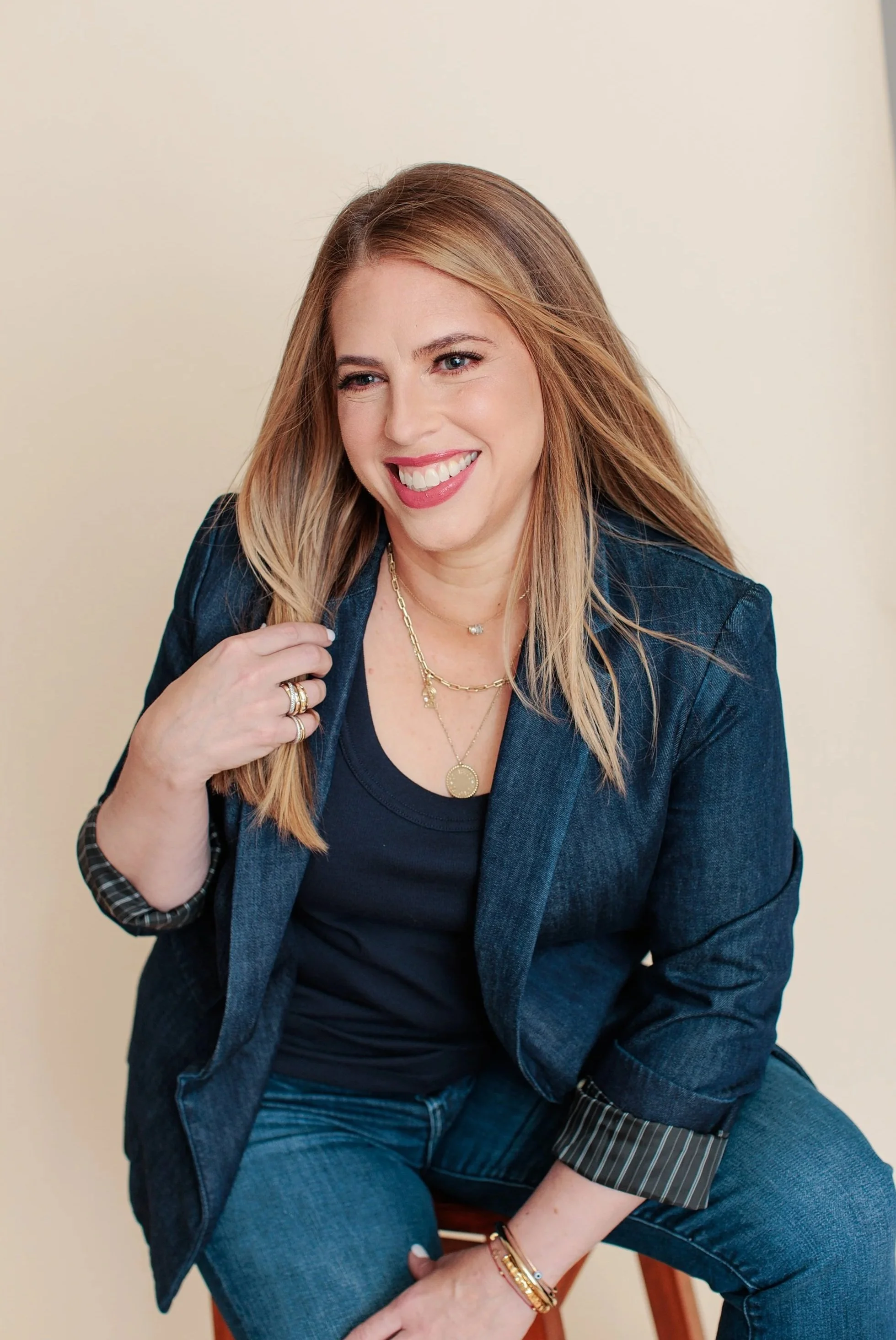 A woman with long blonde hair, wearing a dark jacket and jeans, smiling and sitting on a chair against a plain beige background.