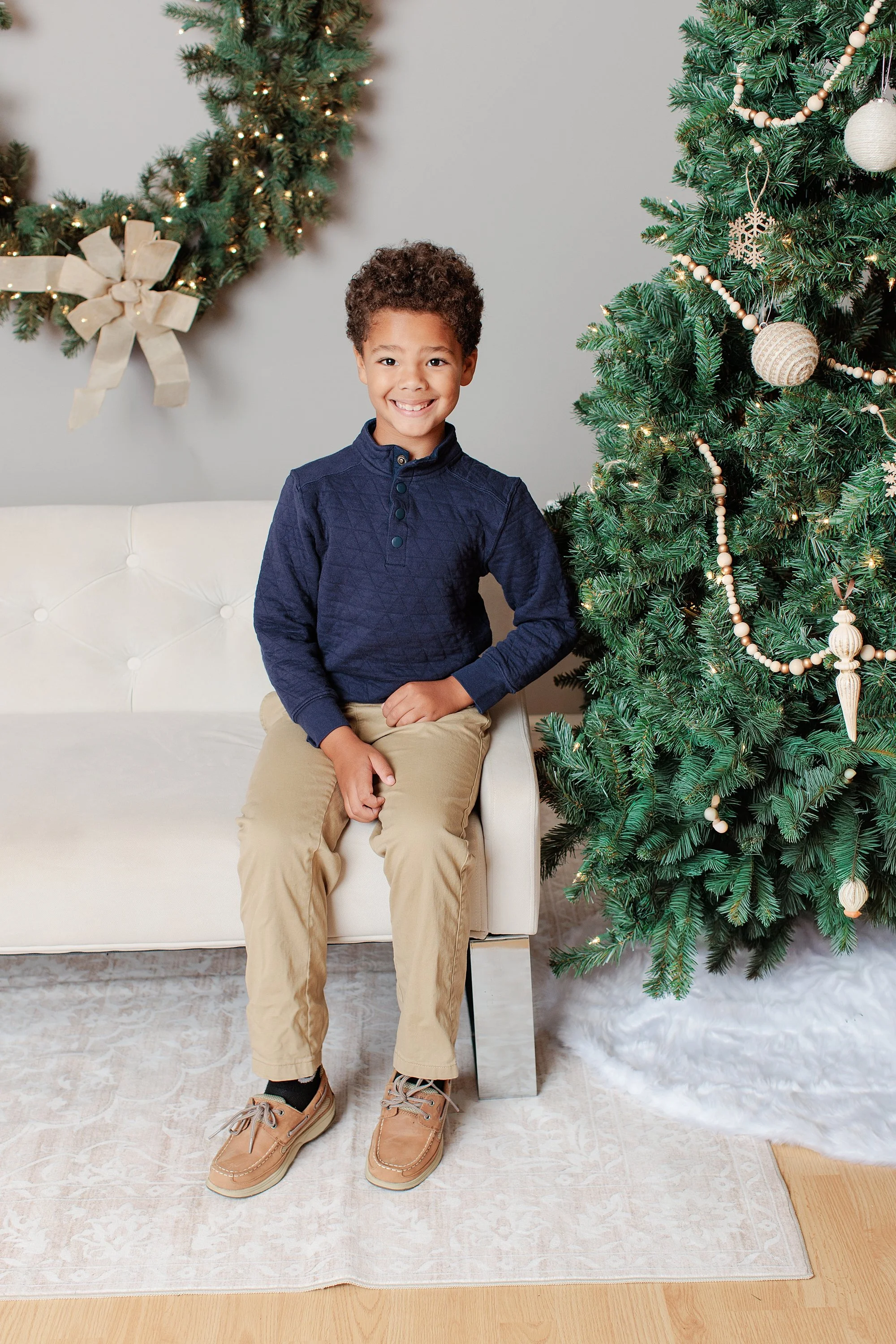 Boy sitting on a cream sofa beside a Christmas tree with neutral ornaments and a lighted wreath.