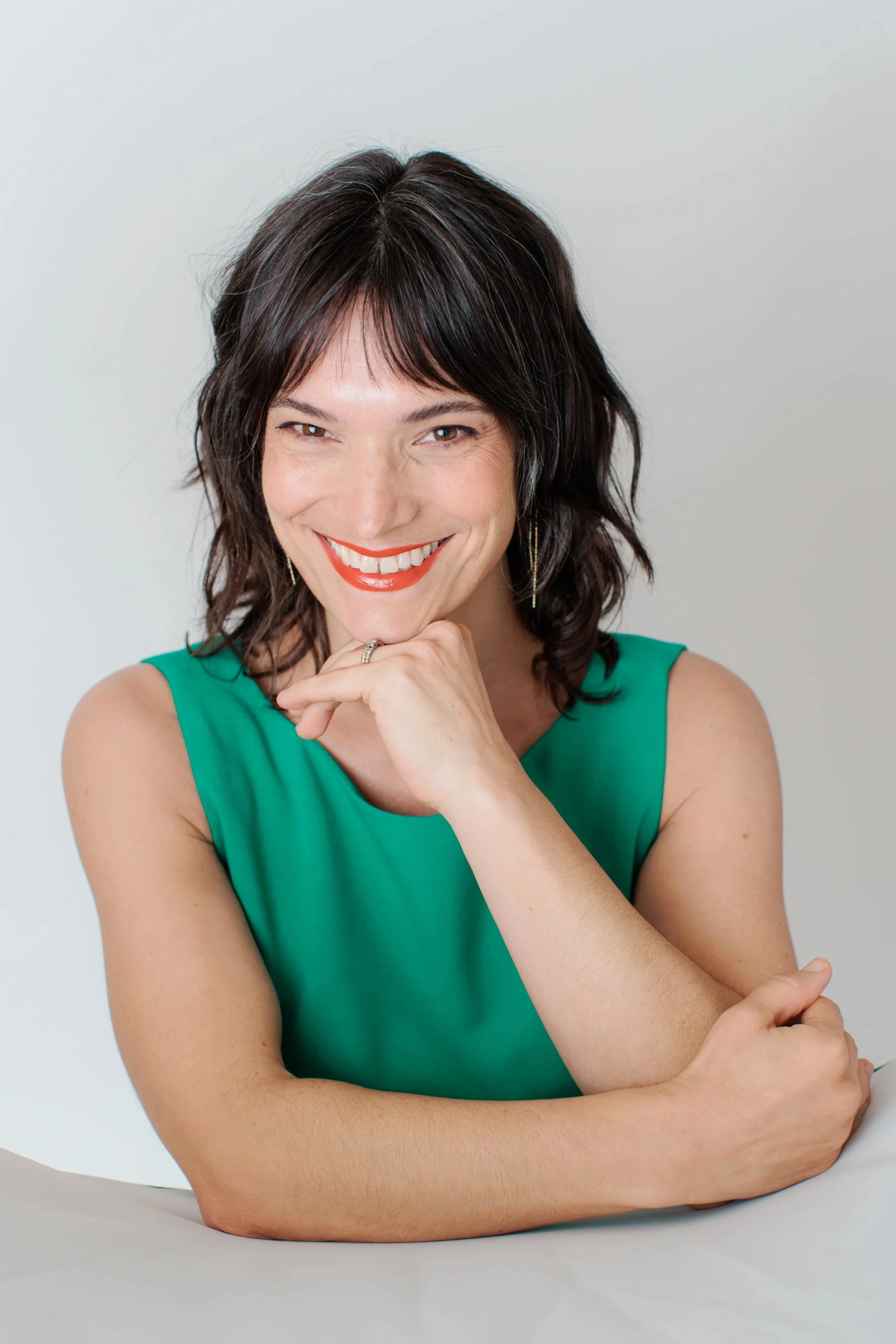 Woman with short dark hair, wearing a green sleeveless dress, smiling with red lipstick, resting her chin on her hand, sitting with arms crossed on a white surface against a plain white background.