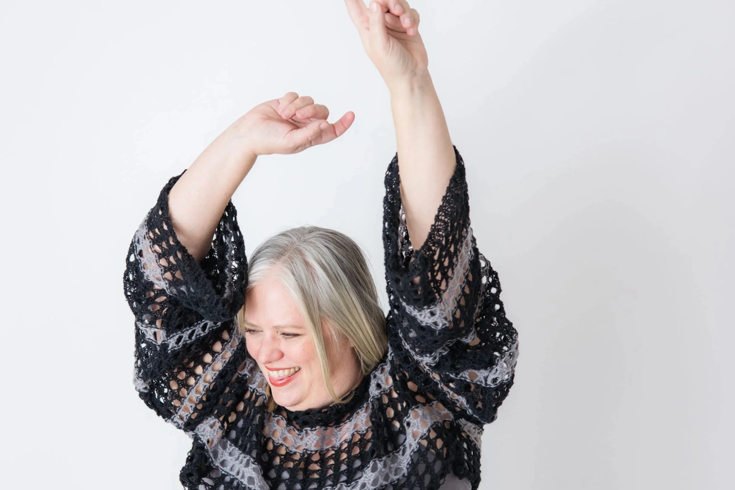An elderly woman smiling with her eyes closed, wearing a black lace dress, with hands raised above her head and her hair showing gray and blonde highlights. The background is plain white.