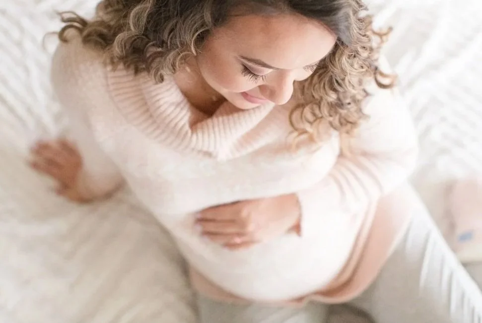 A pregnant woman with curly hair, wearing a cream-colored sweater, sitting on a bed with white bedding, smiling softly and cradling her belly.