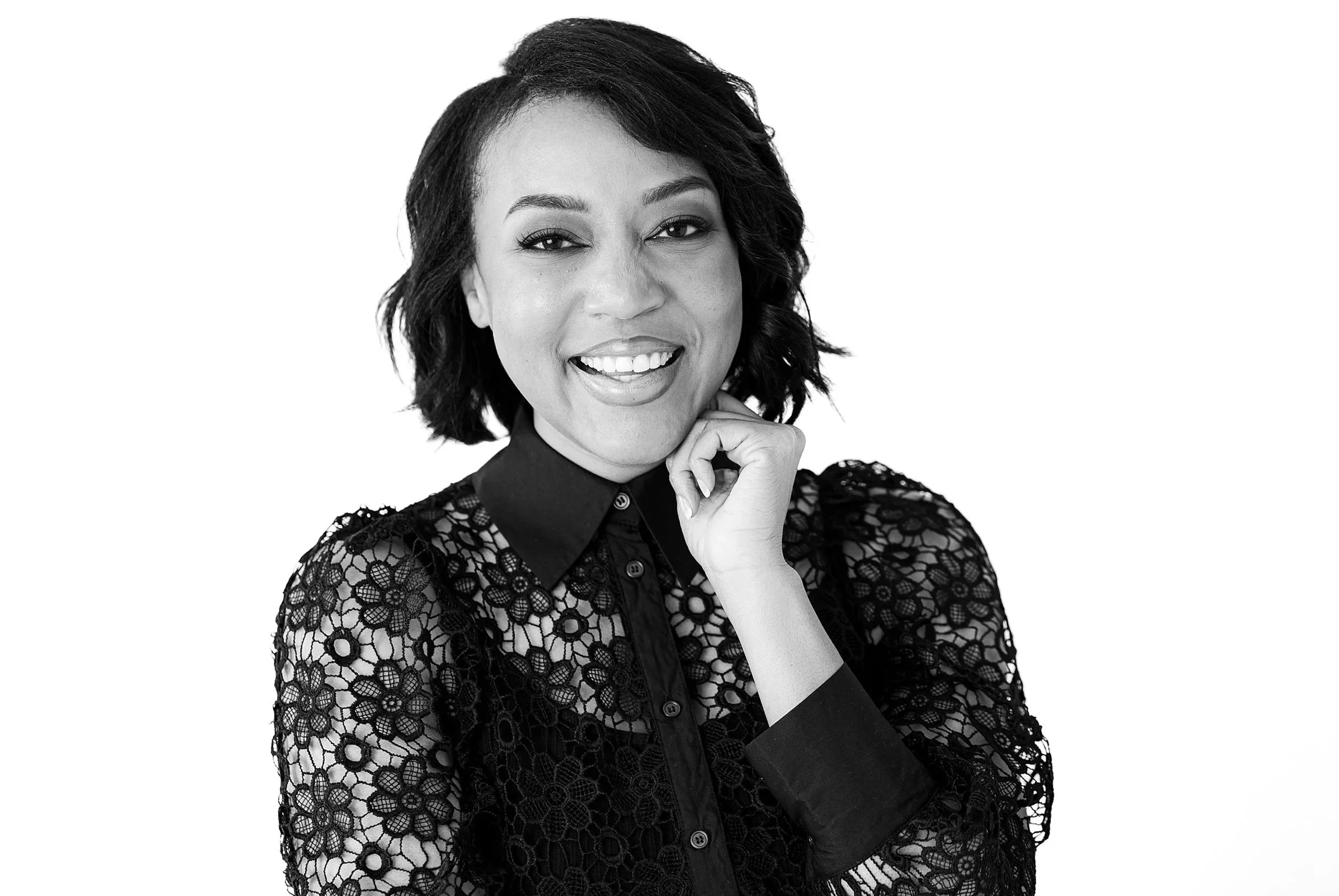A smiling woman with short, wavy hair wearing a lace top and a collared shirt underneath, posing with her hand near her chin against a white background.
