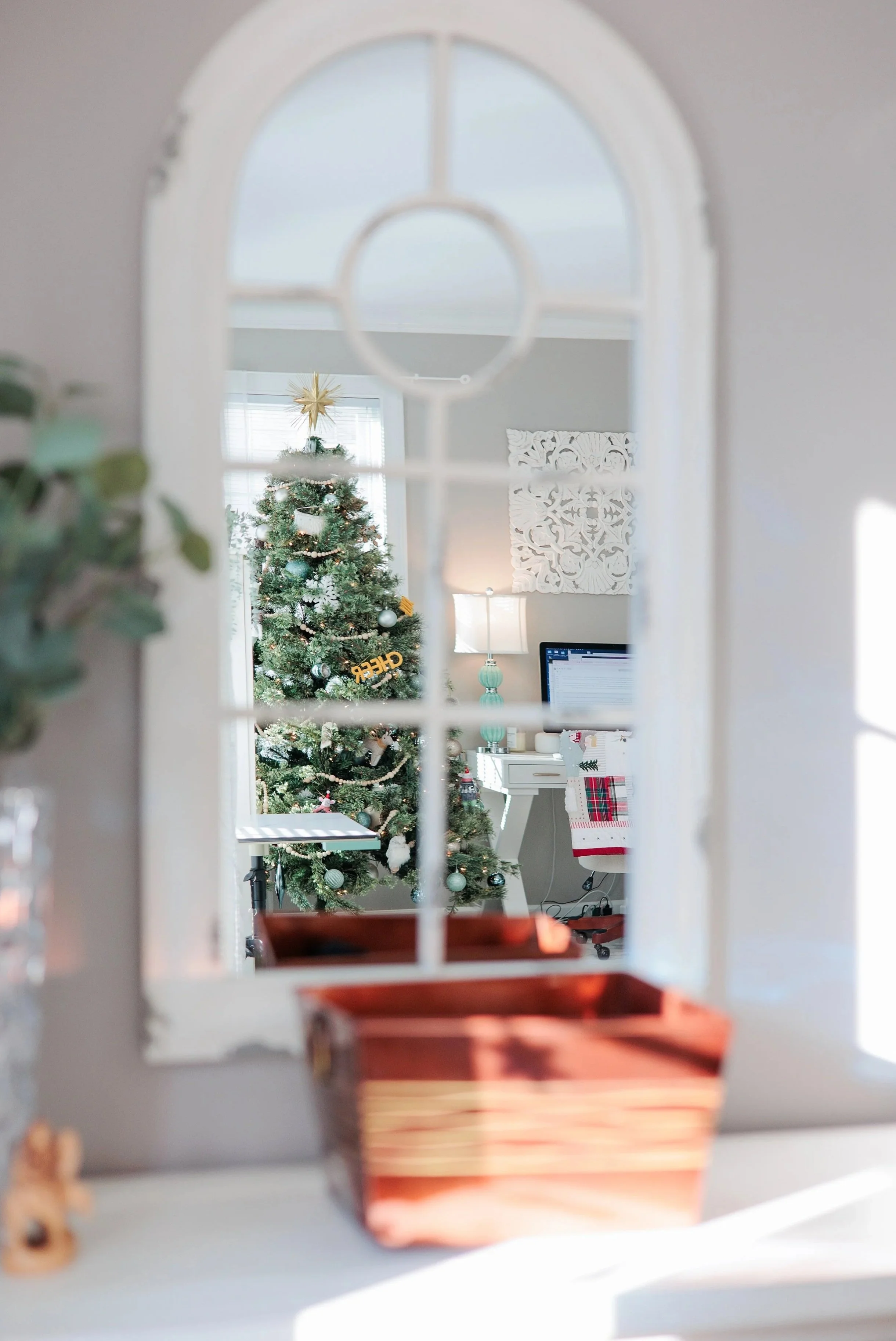 A decorated Christmas tree with ornaments and a gold star topper, viewed through a window with a rounded top and divided panes in a cozy room with white walls, a desk with a computer, and decorative wall art.