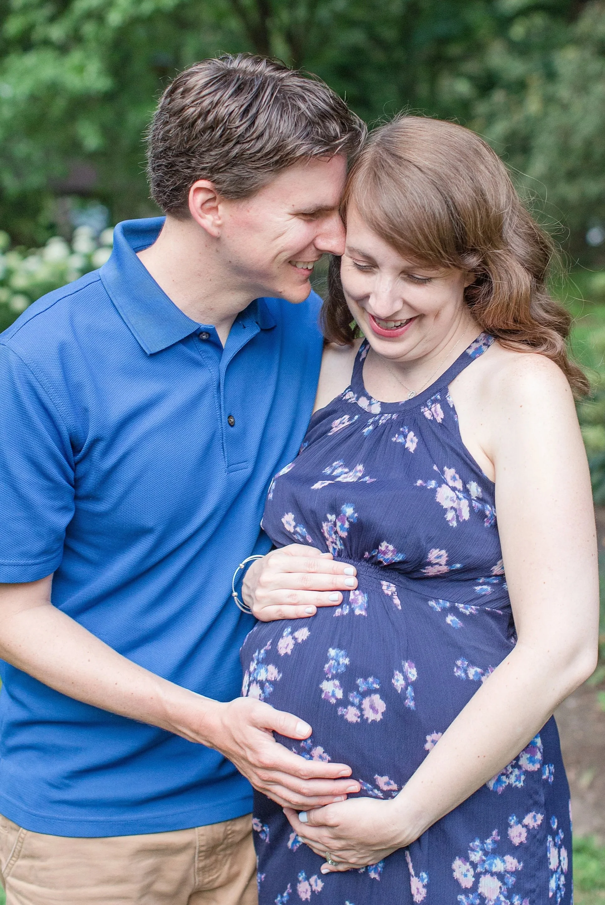 A man and a pregnant woman are smiling and touching her belly together outdoors.