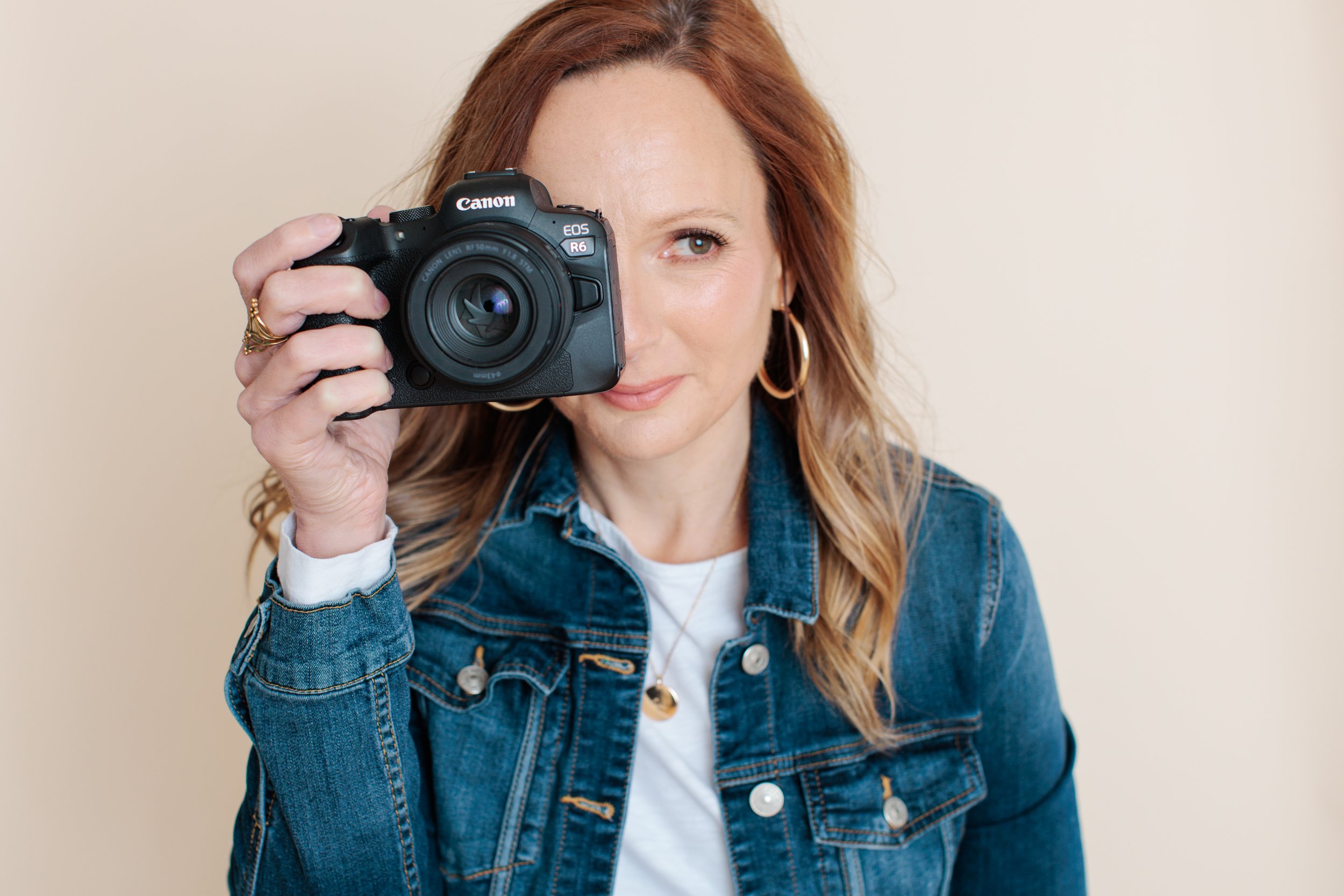 A woman with red hair, wearing a denim jacket and gold hoop earrings, holding a Canon EOS R6 camera close to her face, looking through the viewfinder with one eye.