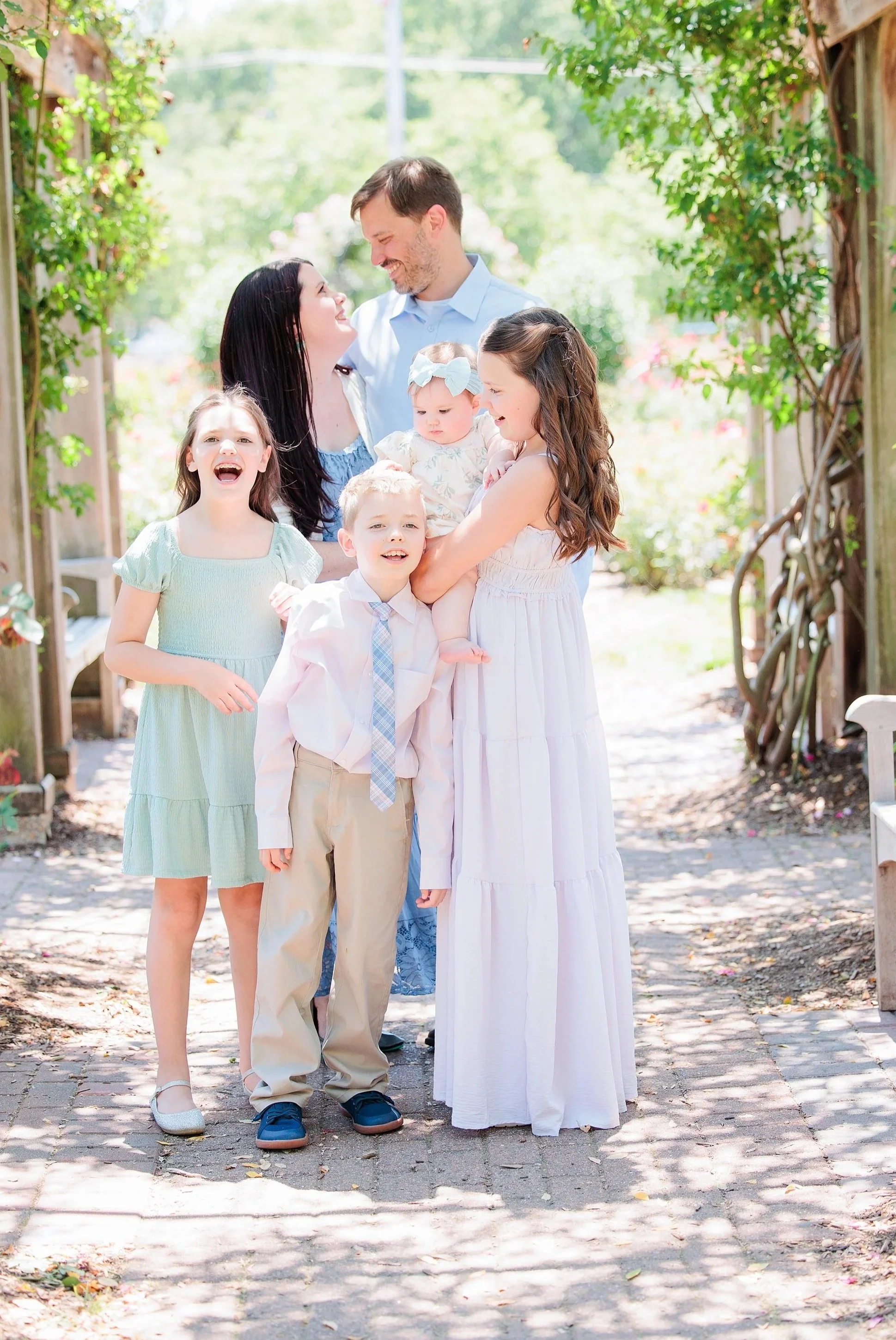 A group of six people, including two adult women, two young girls, a young boy, and a baby, gathered outdoors on a sunny day, smiling and enjoying each other's company.