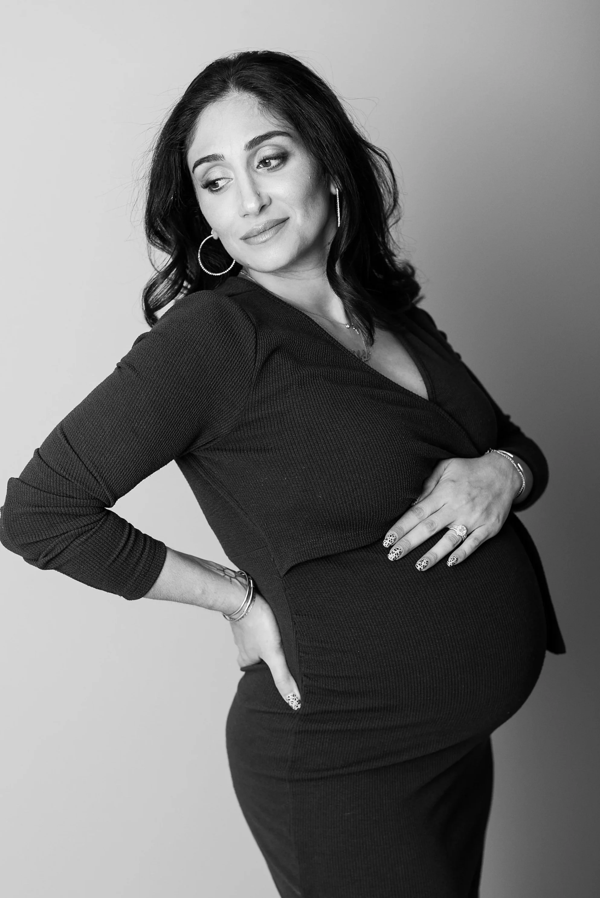 Black and white portrait of a woman with dark hair wearing a fitted dress, wearing hoop earrings and jewelry, standing with one hand on her hip and the other on her belly, looking to her left.
