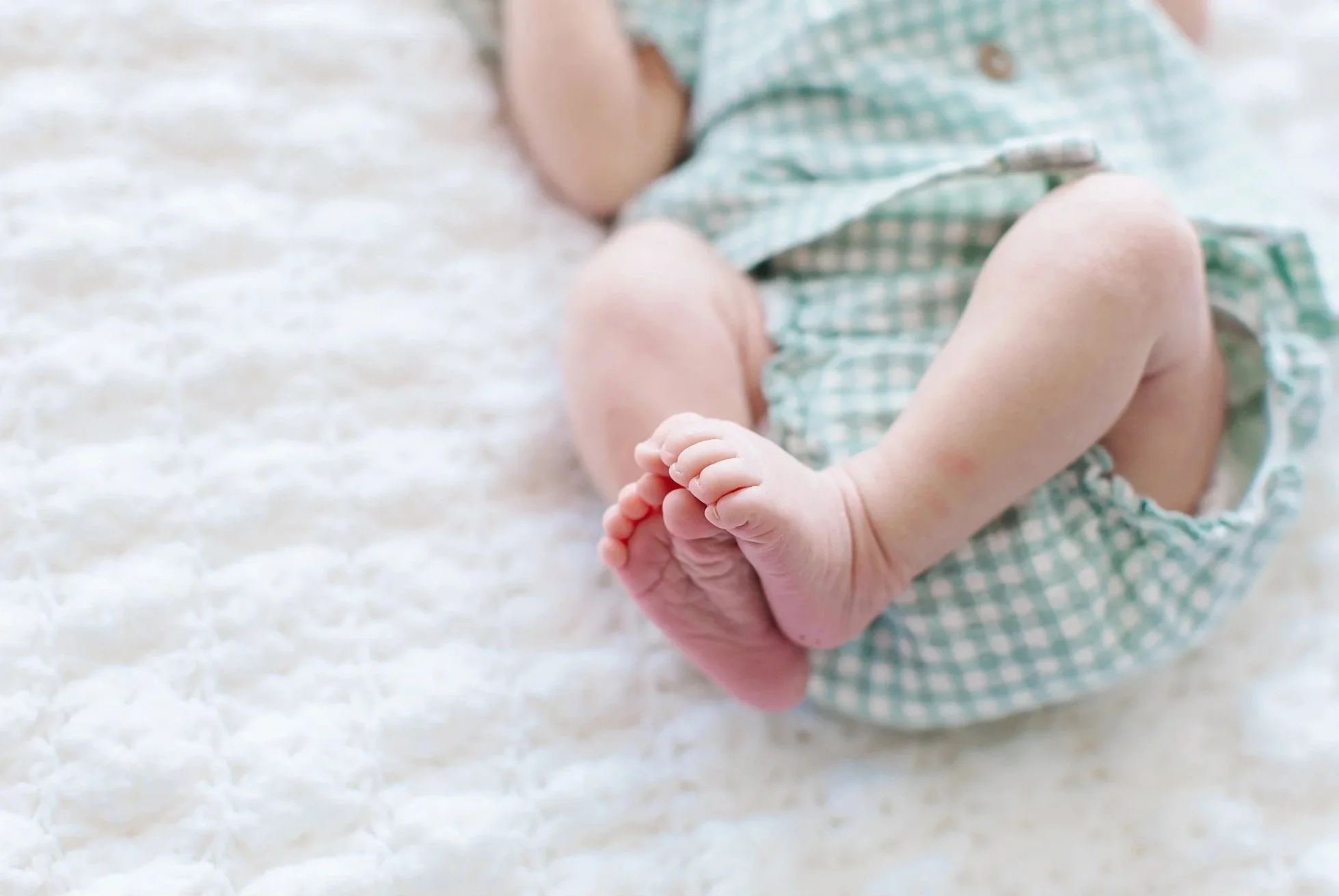 Close-up of a baby's feet and legs, with a blurred background, lying on a soft, white textured surface, wearing a green and white checkered outfit.