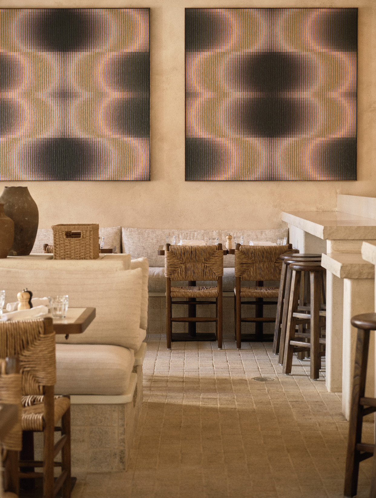 A warm, neural-toned restaurant interior featuring hand-troweled plaster walls, woven rush-seat chairs, and marble tables. Two large, identical abstract painting with vertical stripes and ircular patterns hand side-by-side above a light-colored banq.
