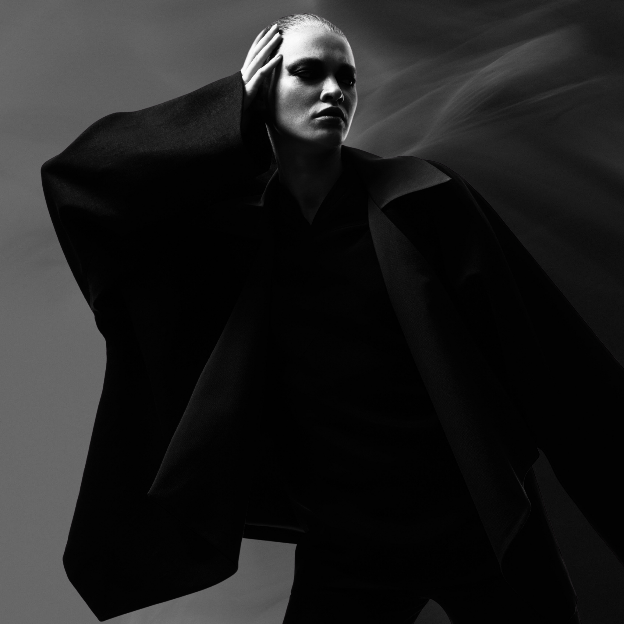 A woman with slicked-back hair dressed in dark clothing poses against a cloudy sky in black and white.